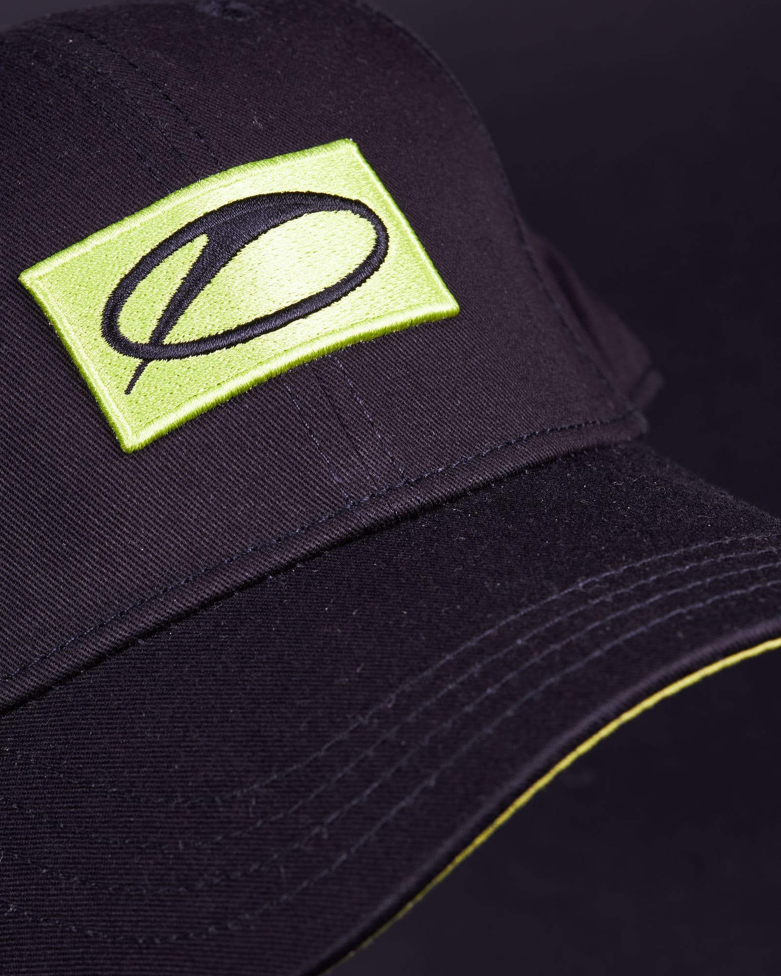 The black cap seen at an angle against a dark ground, with the lime green A State of Trance patch on the front panel and the curve of the stitched brim, edged underneath in lime green.