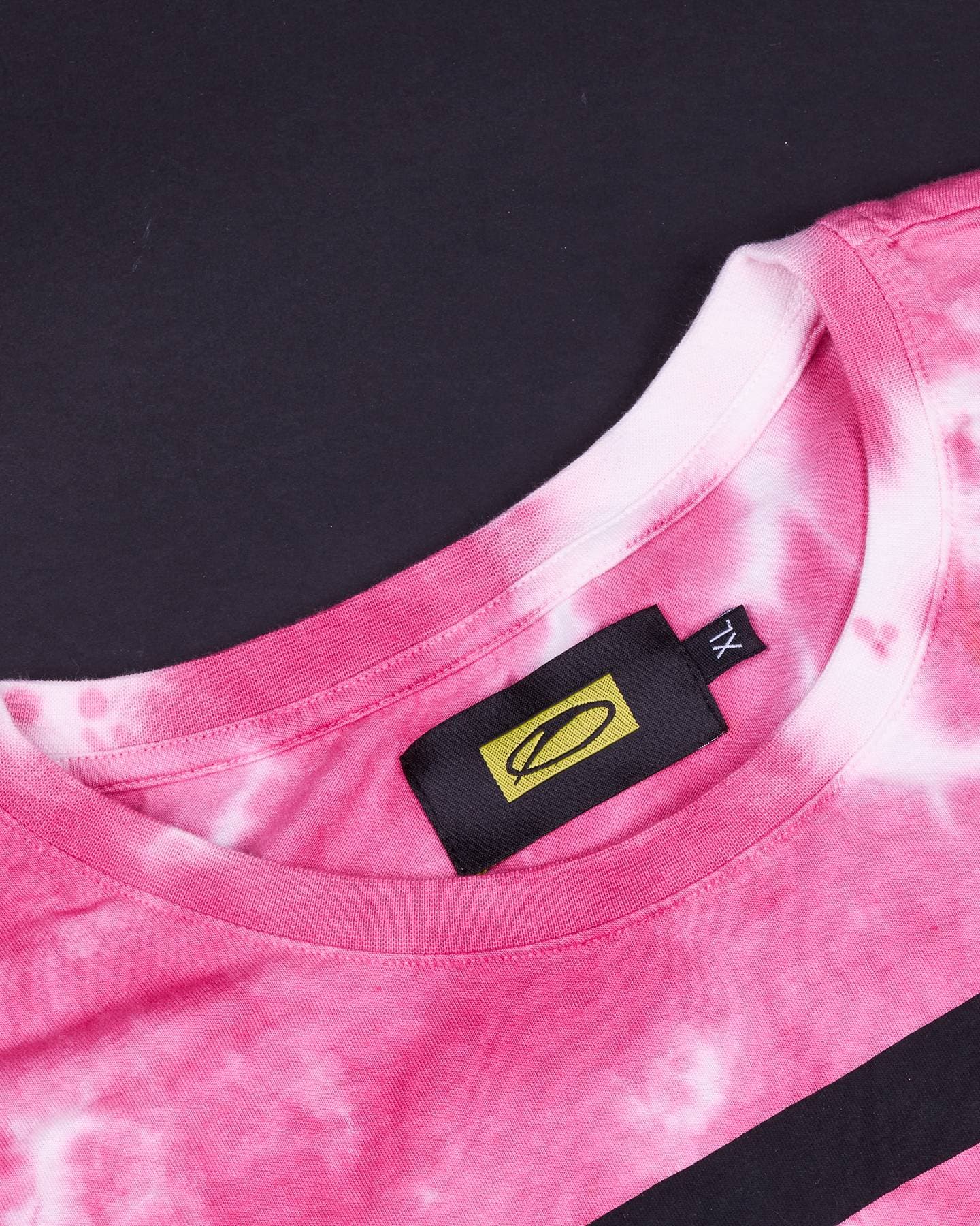 Close-up of the ribbed collar of the pink and white tie-dye T-shirt on a dark ground, with a black woven neck label showing the A State of Trance logo on yellow, a small XL size tab beside it and the edge of the black chest print at the bottom.