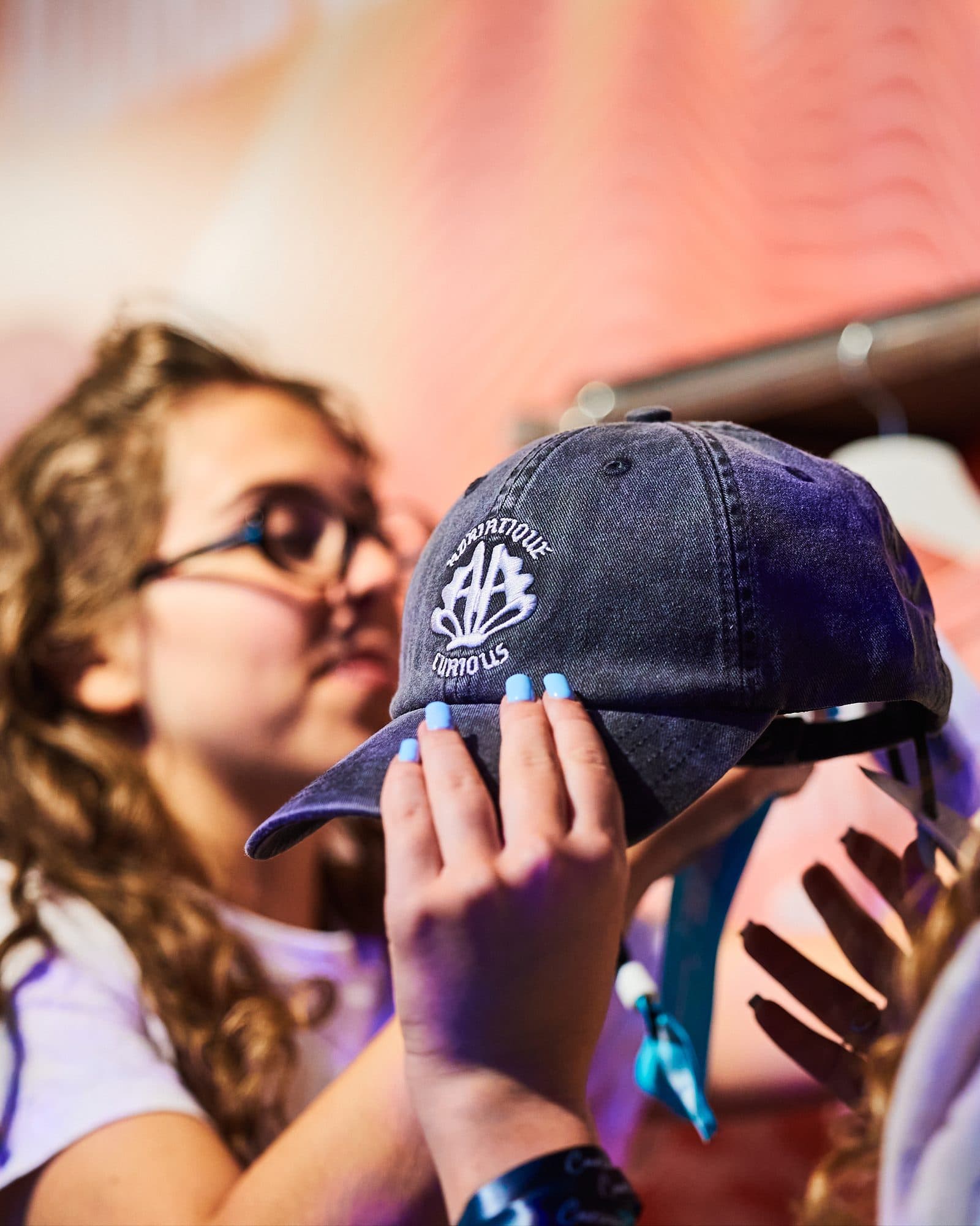 A hand with light blue nails holding up a washed dark grey cap with ADRIATIQUE and CURIOUS embroidered in white around a shell-shaped monogram, in front of a person in glasses with long curly hair.