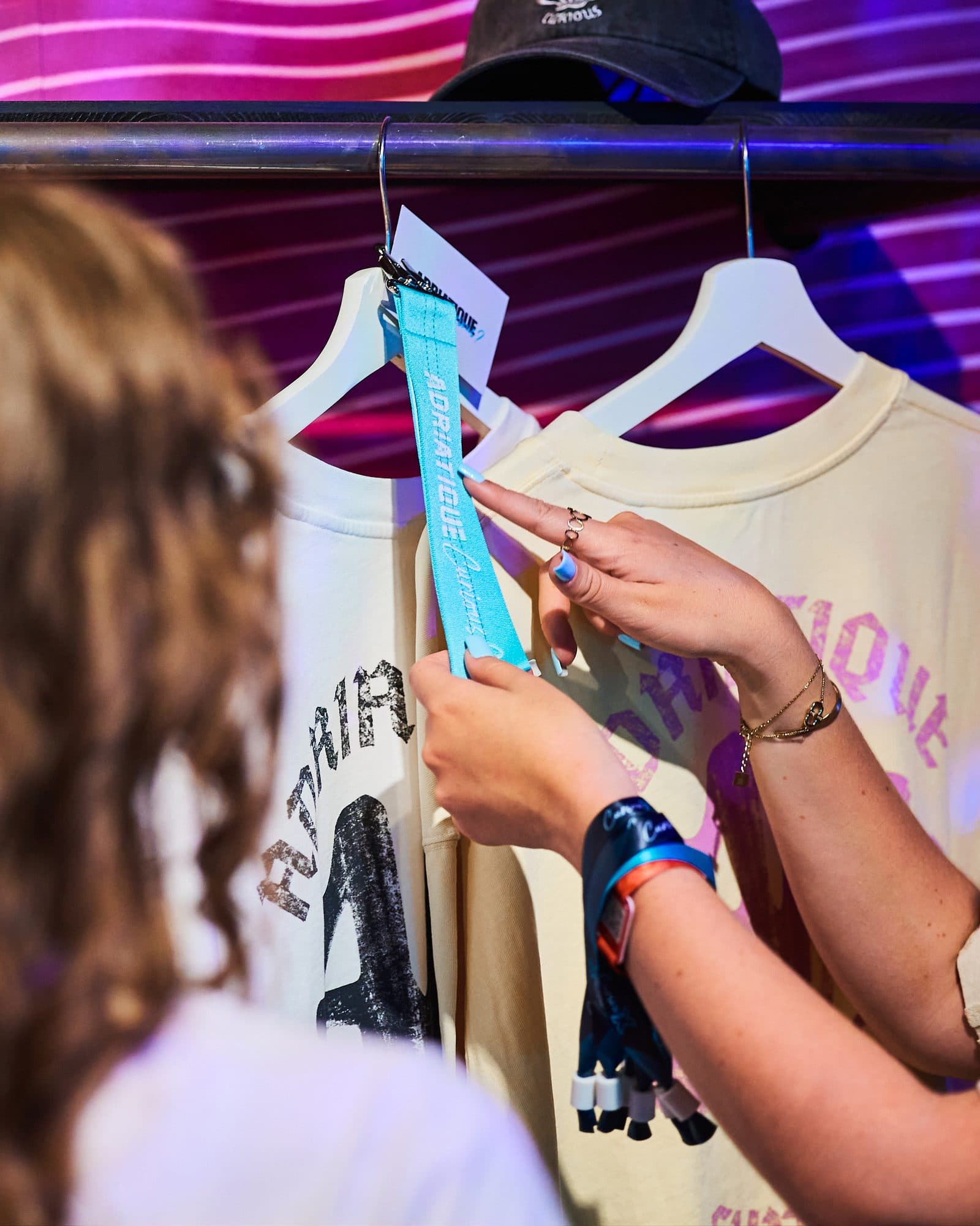 Two hands with light blue nails holding up a teal lanyard with ADRIATIQUE and Curious in white, clipped to a hanger, in front of two cream T-shirts with black and pink Adriatique prints, a dark grey cap on the rail above and a person with long hair in the foreground.