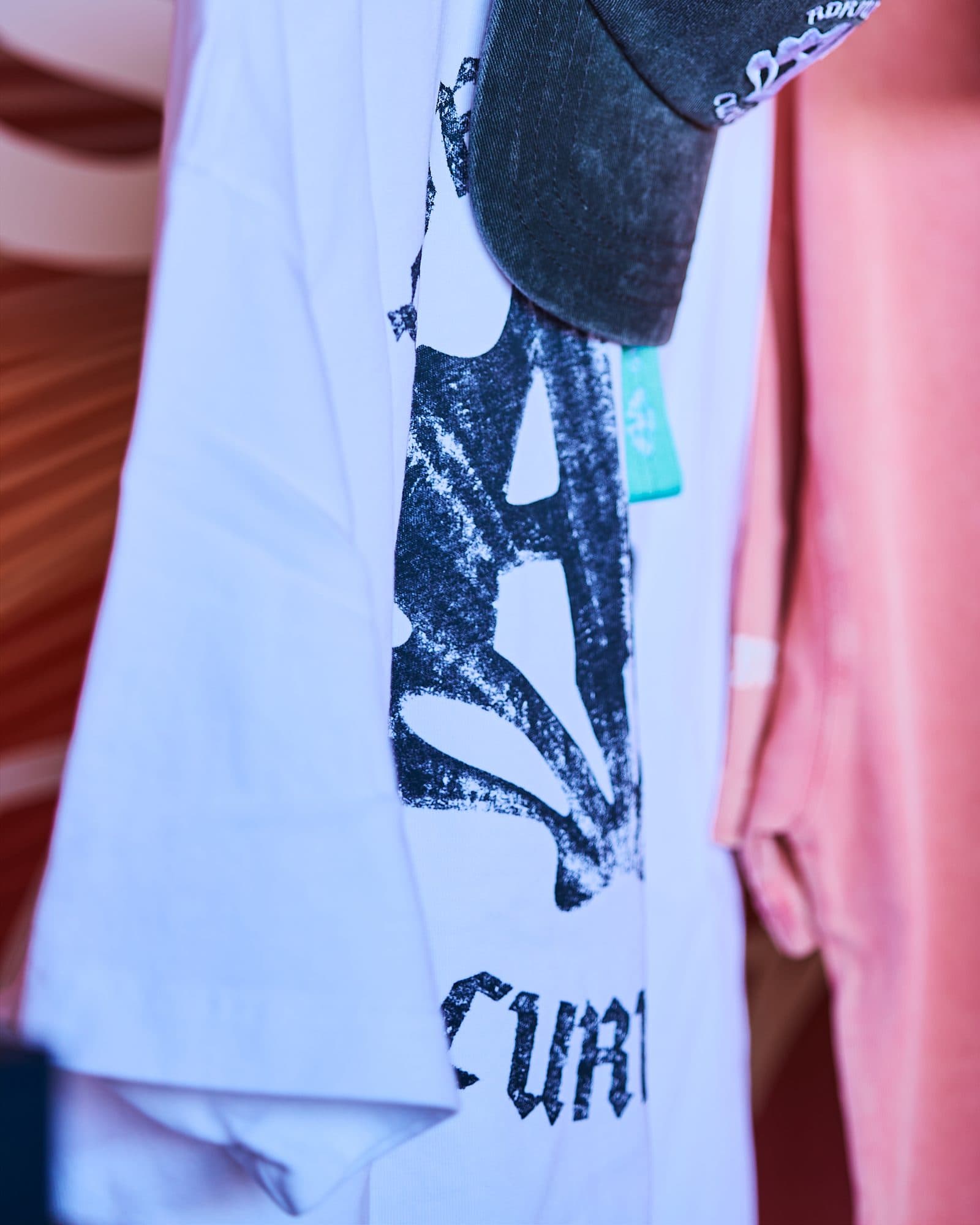 A white T-shirt with a large distressed black print of a shell-shaped monogram above the first letters of CURIOUS, under the brim of a washed dark grey cap, with a small teal label and a pink T-shirt beside it.