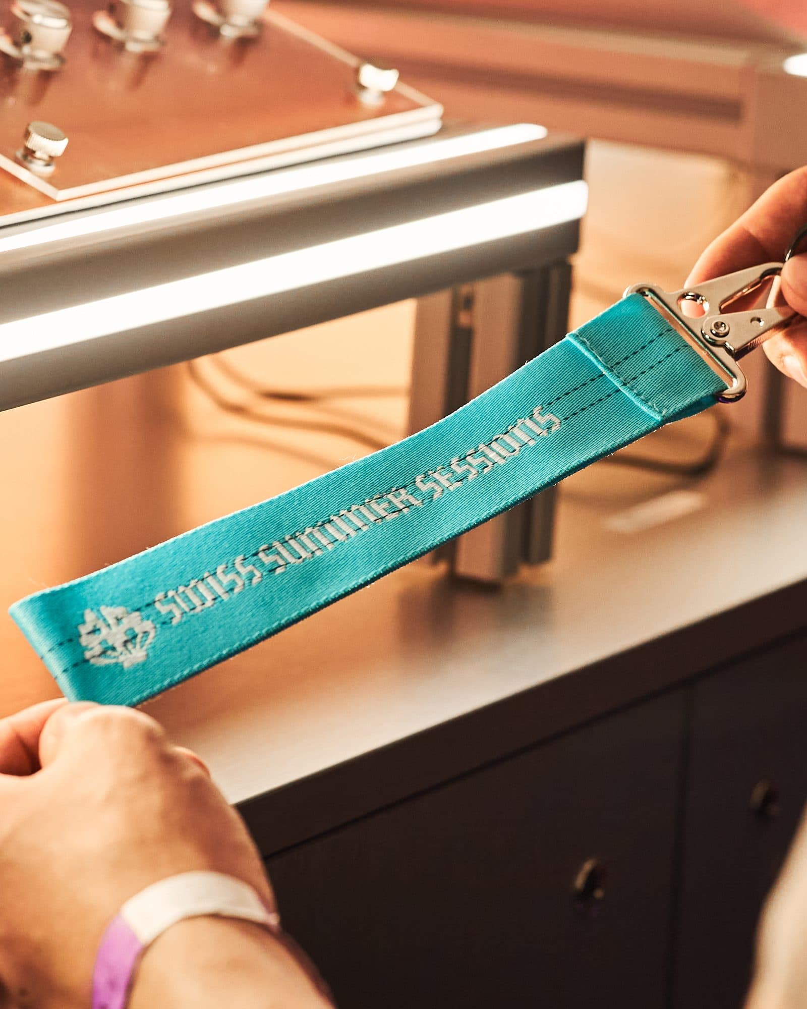 Two hands holding a short teal lanyard with a metal snap hook, with a small emblem and SWISS SUMMER SESSIONS in white, over a lit counter with metal knobs.