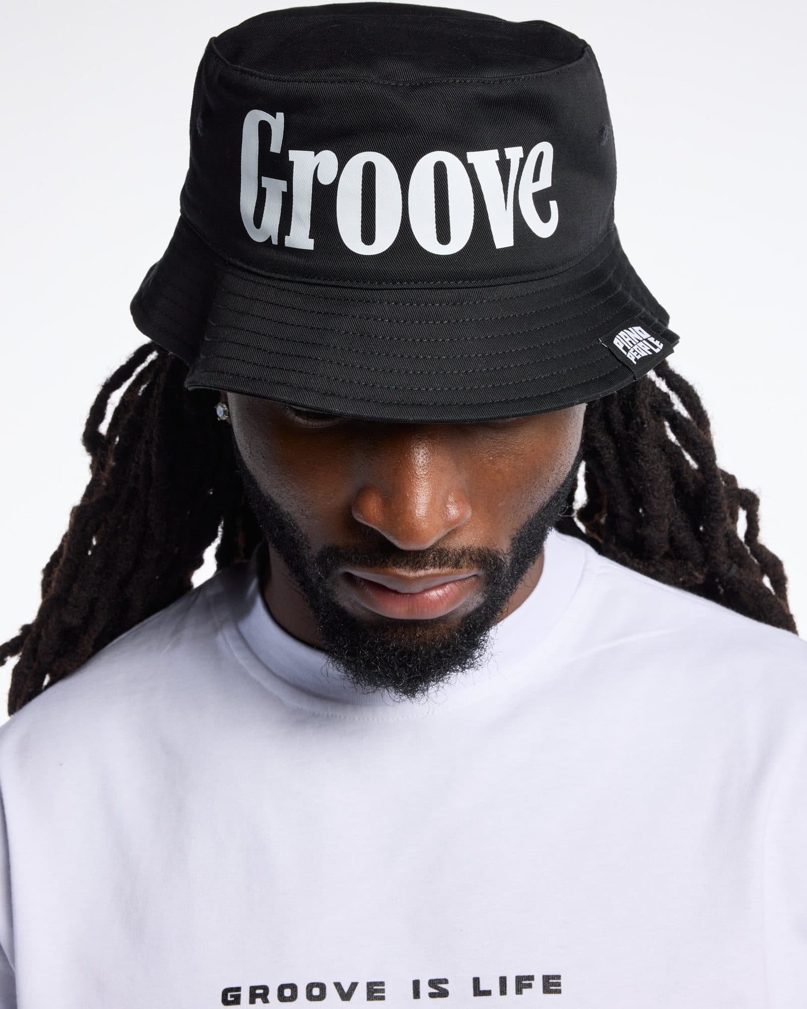 A person with long locs looking down, in a black bucket hat printed with Groove in large white letters and a small PIANO PEOPLE label on the brim, and a white T-shirt with GROOVE IS LIFE in black on the chest.