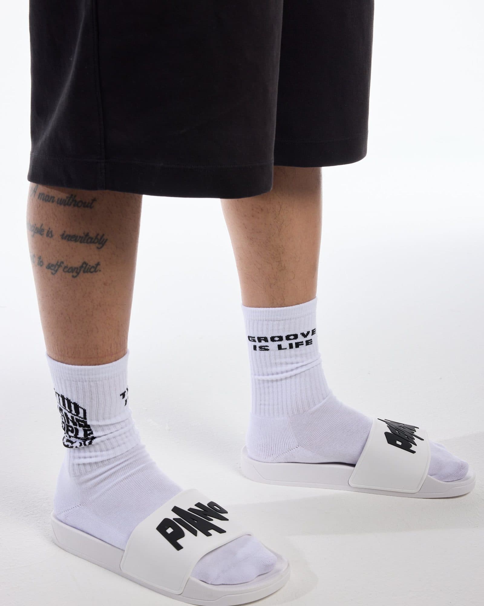 Legs below black shorts, in white ribbed socks, one with GROOVE IS LIFE in black and the other with a black logo partly turned away, and white sliders with PIANO in large black letters on the strap, on a white floor.