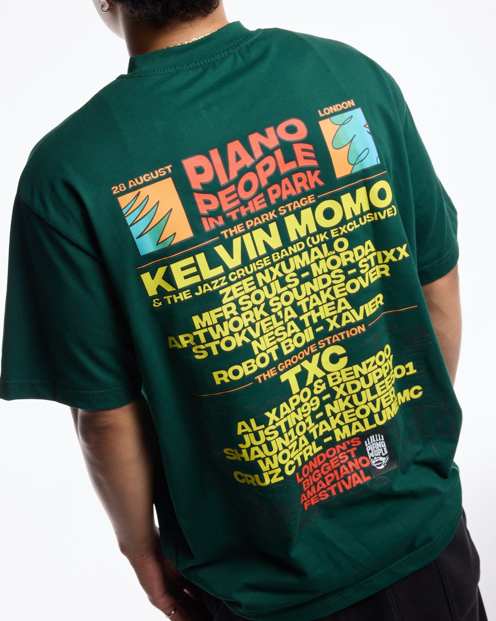 A person seen from behind against a white background, in a dark green T-shirt with a back print: PIANO PEOPLE IN THE PARK in red, 28 AUGUST and LONDON beside two square graphics, a line-up in yellow letters, and LONDON'S BIGGEST AMAPIANO FESTIVAL in red beside a PIANO PEOPLE badge.