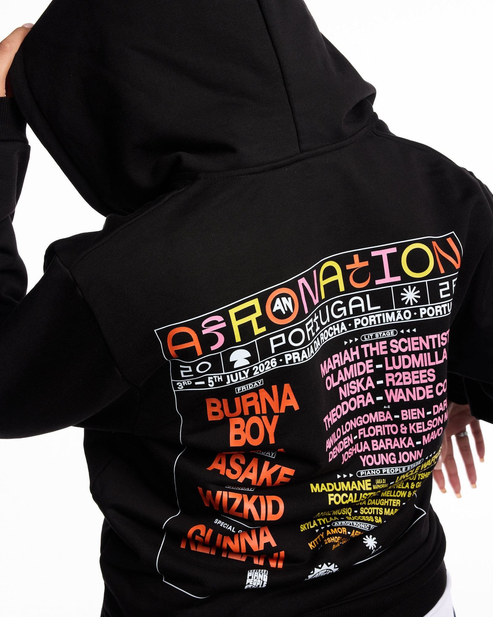 A person seen from behind against a white background, one hand on the raised hood of a black hoodie whose back is printed with AFRO NATION in multicoloured letters, PORTUGAL and July 2026 dates in white, and a line-up in orange, pink and yellow inside a white frame.