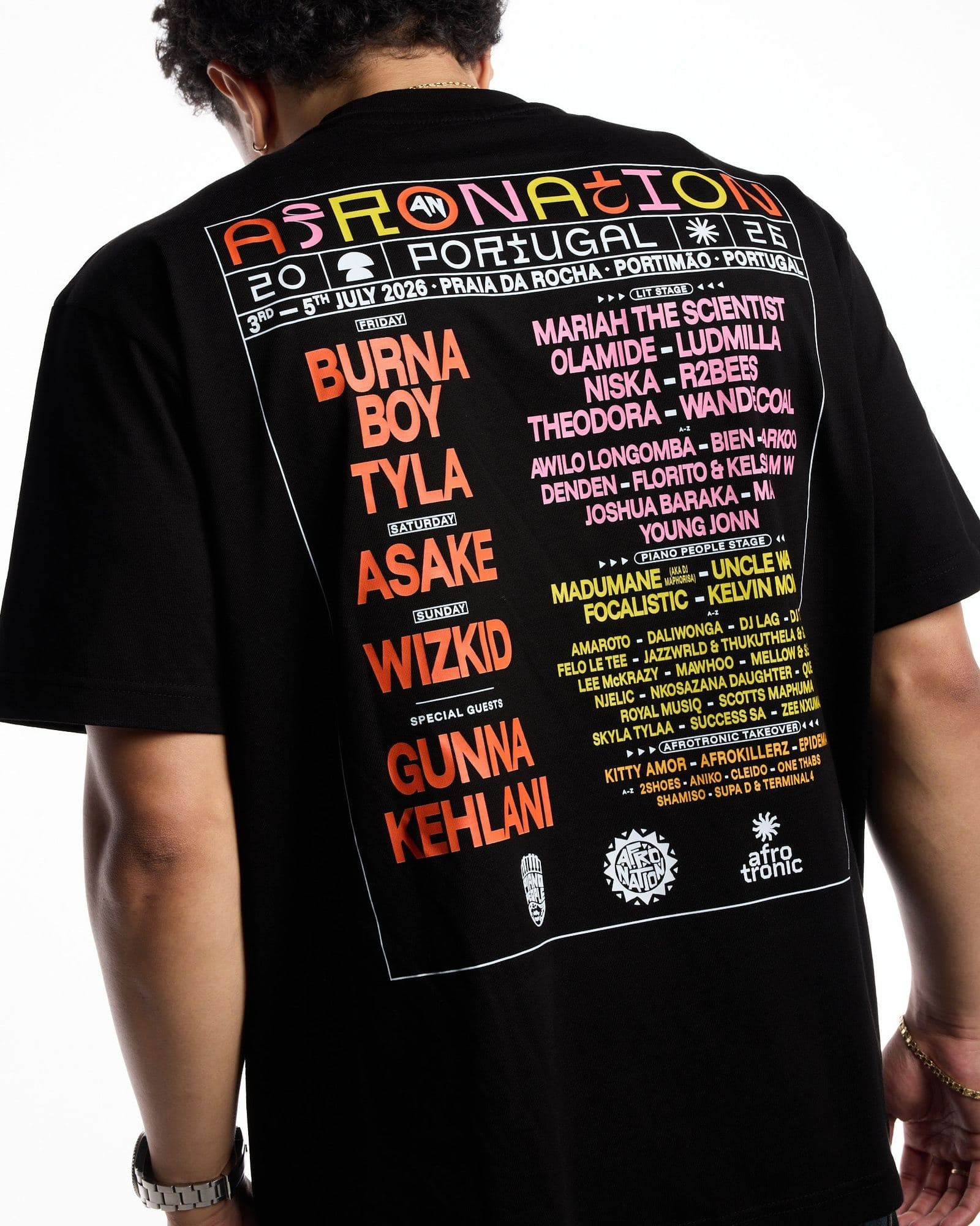 A person seen from behind with head bowed against a white background, in a black T-shirt with a large back print: AFRO NATION in multicoloured letters, PORTUGAL 26, PRAIA DA ROCHA, PORTIMÃO, PORTUGAL and July 2026 dates in white, and a line-up in orange, pink and yellow letters inside a white frame.