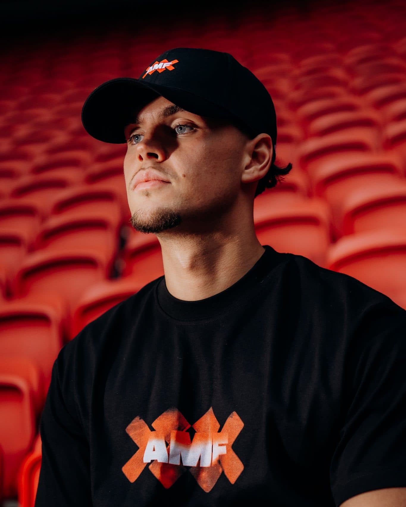 A person in front of rows of red stadium seats, in a black cap with a small AMF and orange-red crosses logo on the front and a black T-shirt with AMF in white letters over large orange-red crosses on the chest.