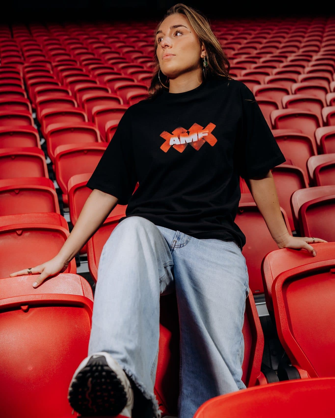 A person with long hair sitting back among rows of red stadium seats, one foot raised towards the camera, in a black T-shirt with AMF in white letters over orange-red crosses on the chest and wide light blue jeans.