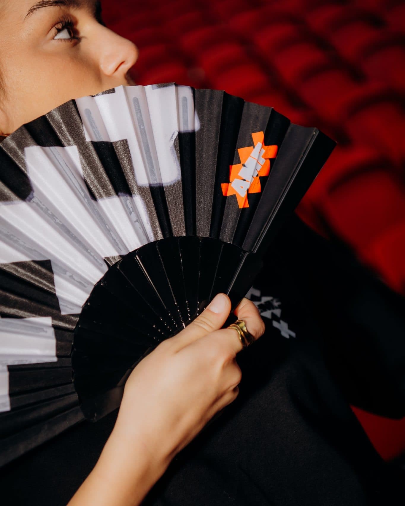 A hand with a gold ring holding an open black, grey and white folding fan in front of the lower half of a person's face, the fan printed with AMF in white letters on orange crosses, red seats behind.