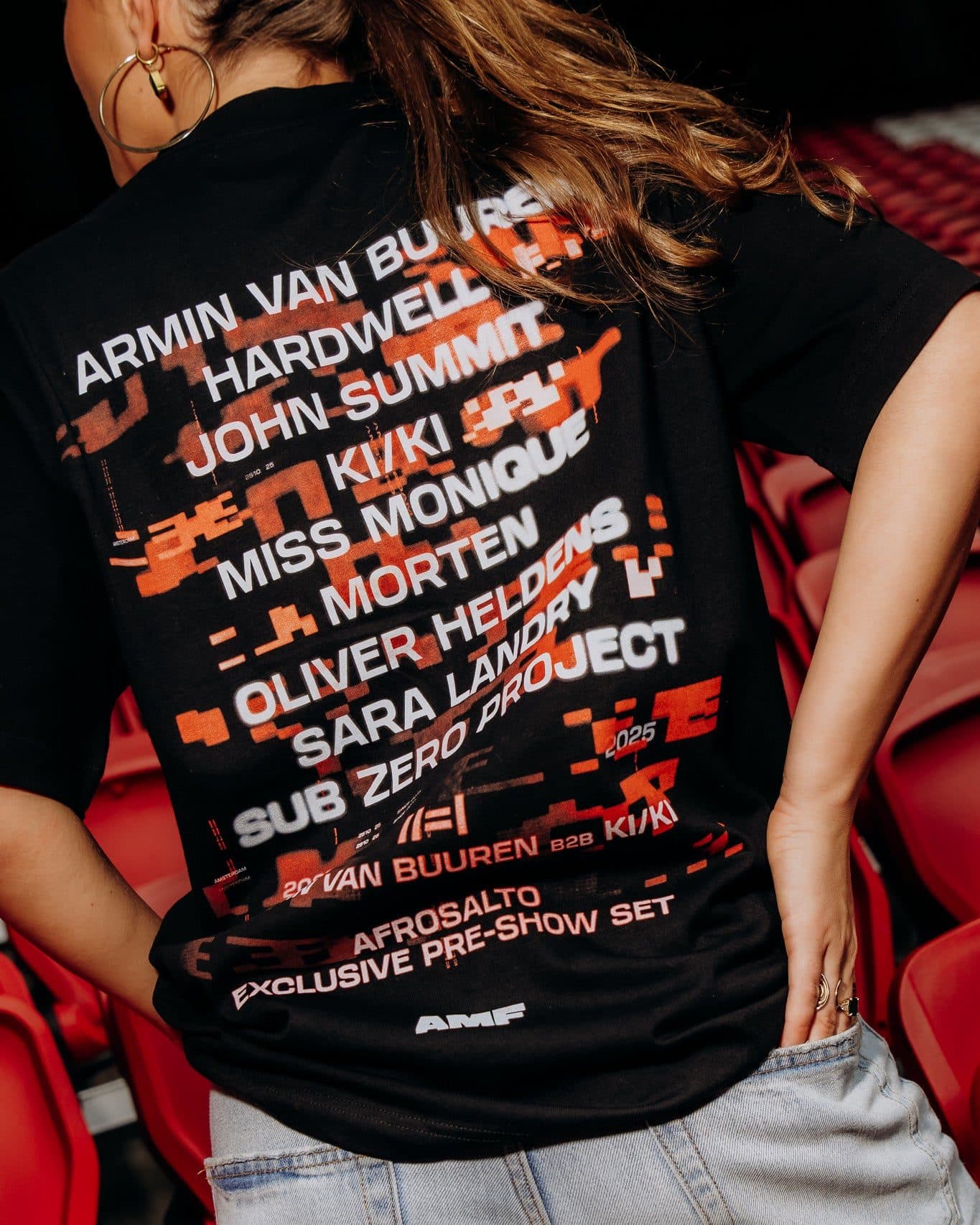 Back of a black T-shirt worn by a person with long hair among red stadium seats, one hand in the back pocket of light blue jeans, printed with a long list of names in large white letters over orange pixel shapes, with 2025 small among the names and AMF near the hem.