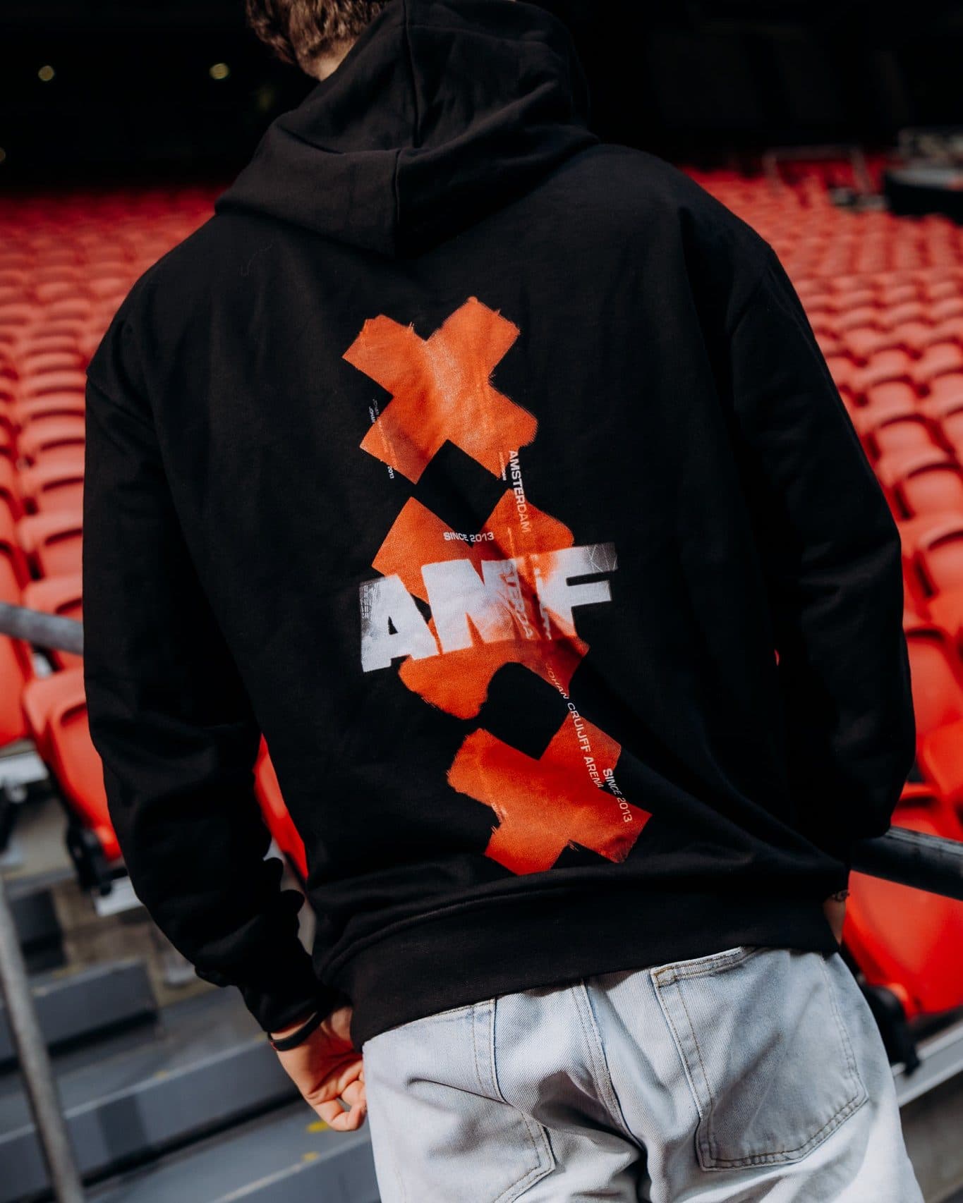 A person seen from behind on the steps of a stadium stand with rows of red seats, in a black hoodie with three orange-red crosses down the back, AMF in large white letters across the middle one and small white lettering beside them, worn with light blue jeans.