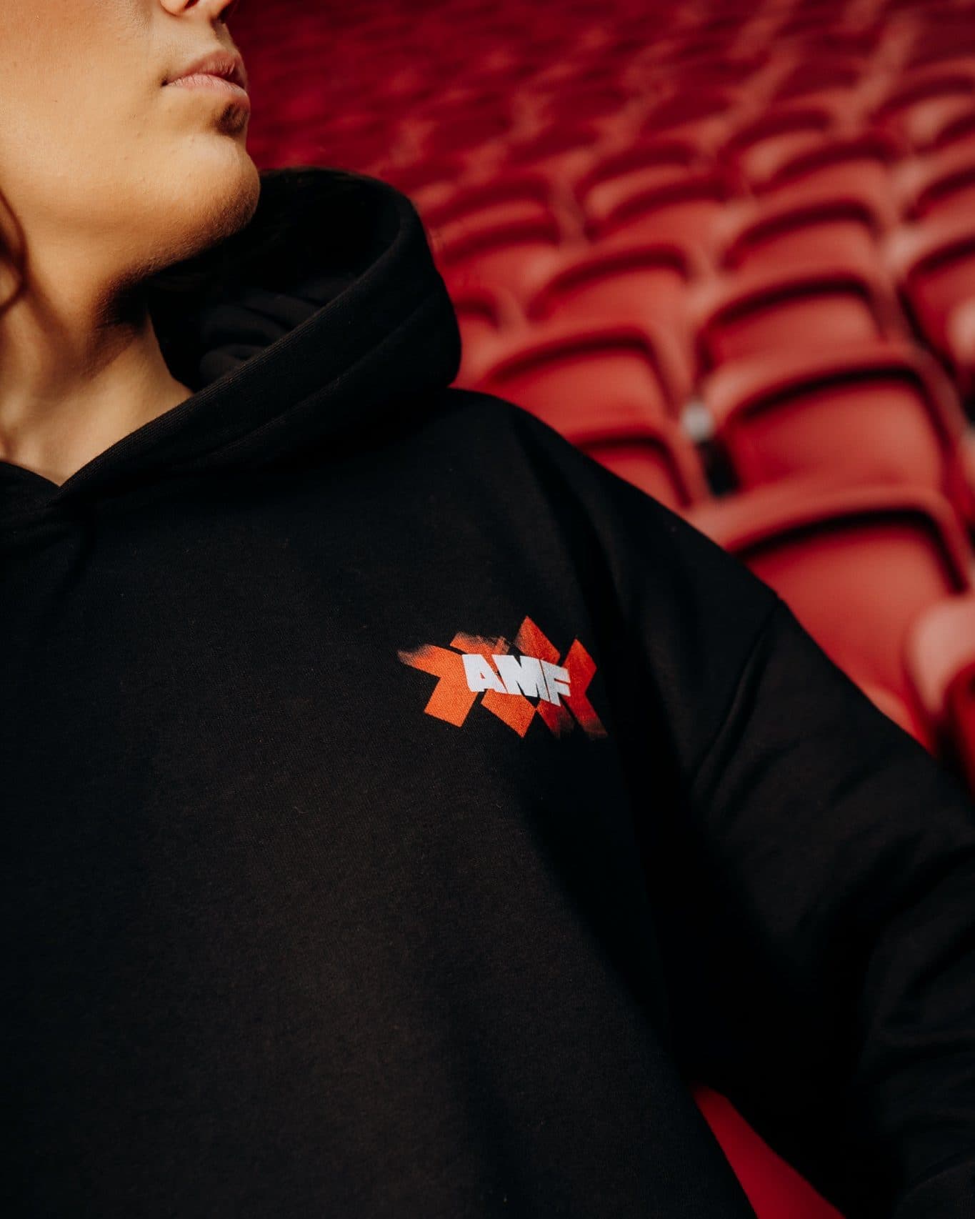 Close-up of the chest of a black hoodie worn by a person in front of rows of red stadium seats, with a small print of orange-red crosses and AMF in white letters.