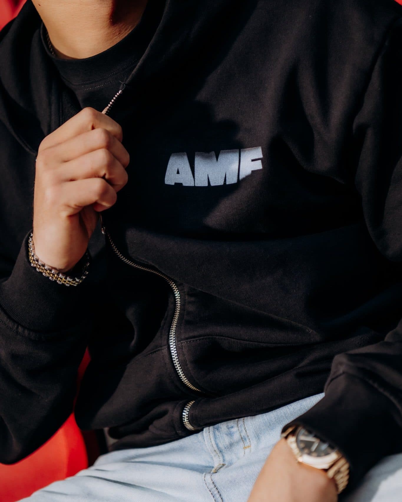 Close-up of a black zip-up worn over a black T-shirt, with AMF in white letters on the chest, one hand holding the silver zip and the other, with a gold watch, resting on light blue jeans.