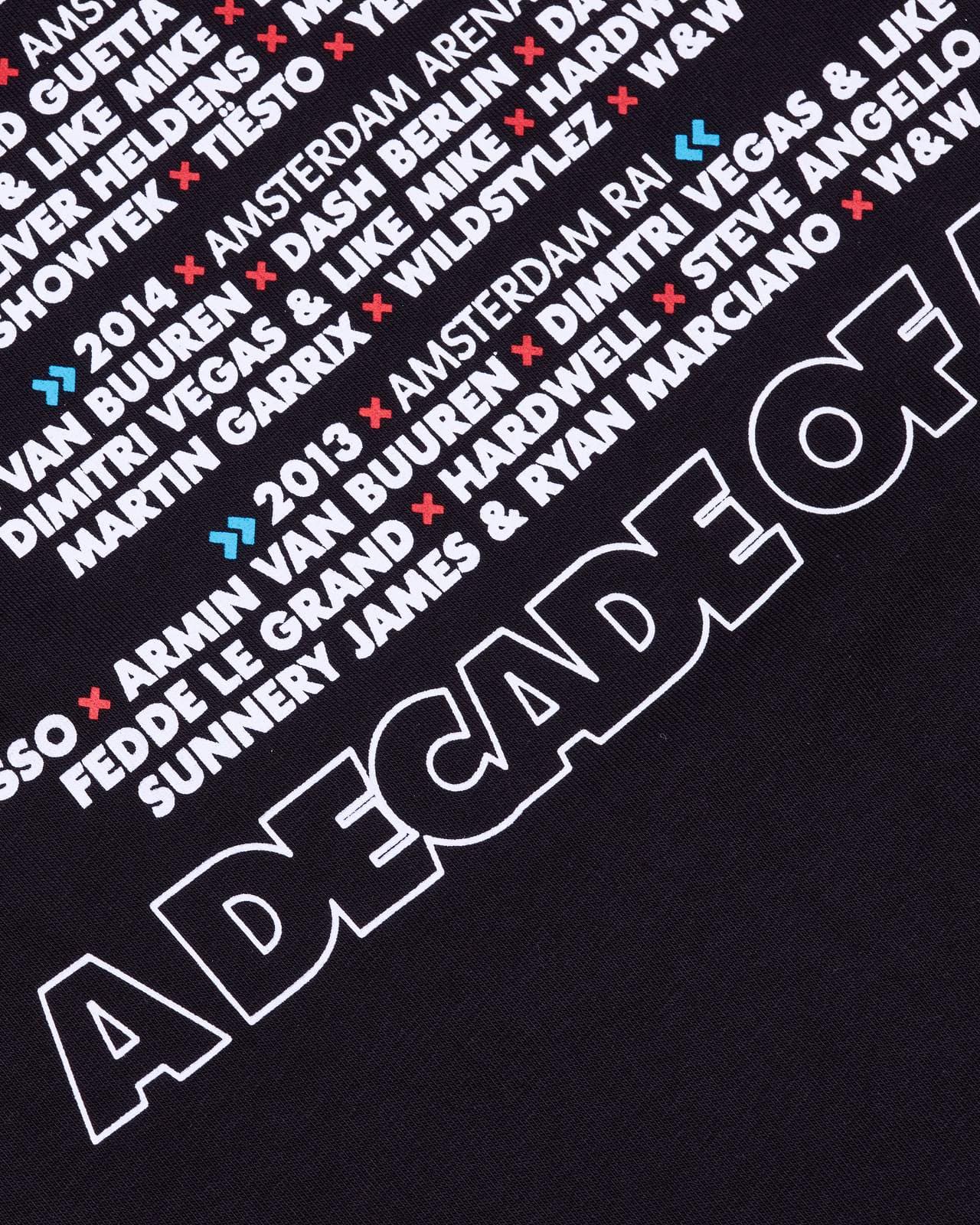 Close-up of the back of a black T-shirt printed with diagonal rows of names in white capitals separated by small red crosses, the years 2013 and 2014 with place names in thinner letters between small blue arrows, and A DECADE OF in large white outlined letters running off the edge.