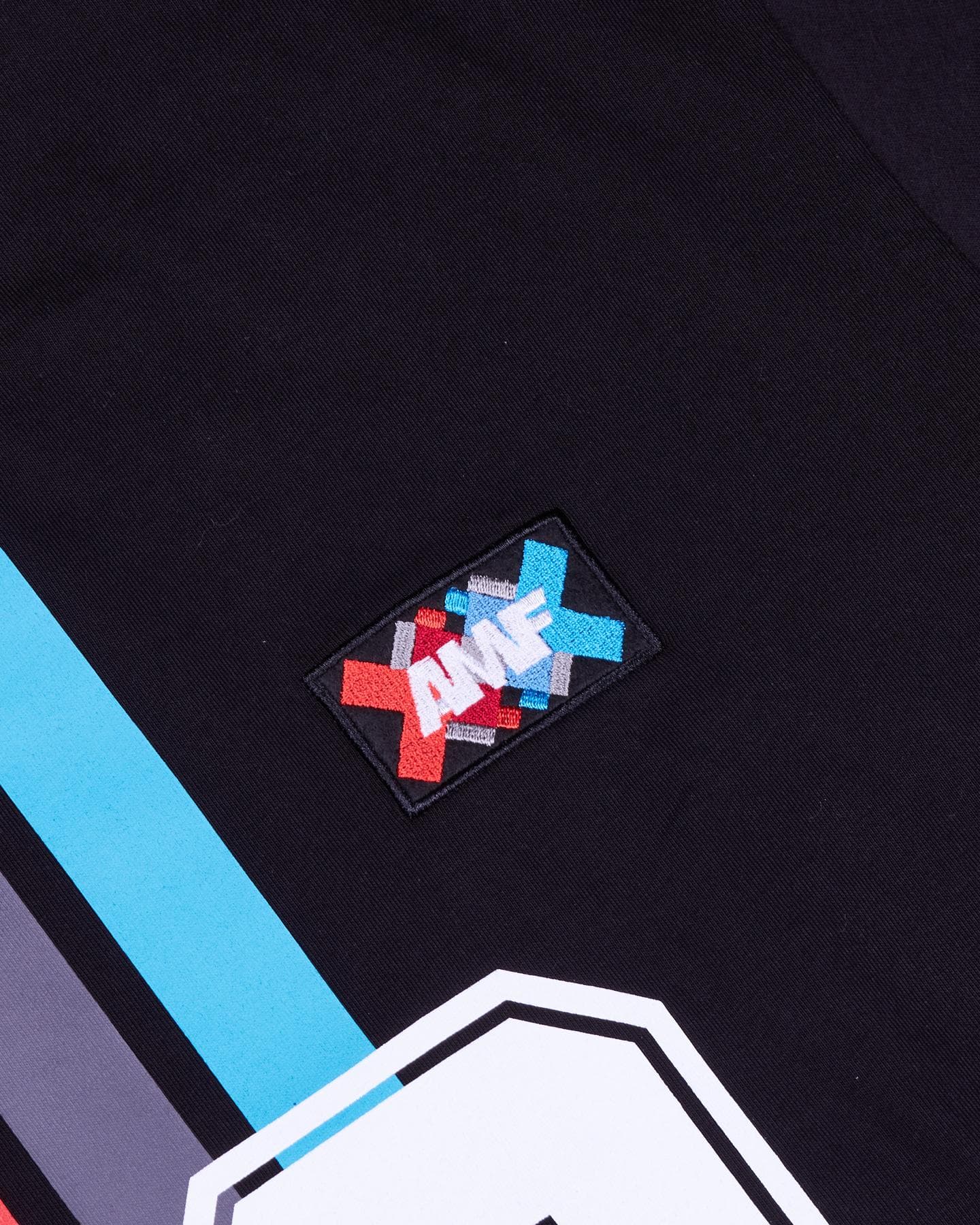Close-up of a small embroidered patch with AMF in white over red, grey and blue crosses on the chest of a black T-shirt, beside blue and grey stripes running diagonally and the top of a large white shape edged in black at the bottom edge.