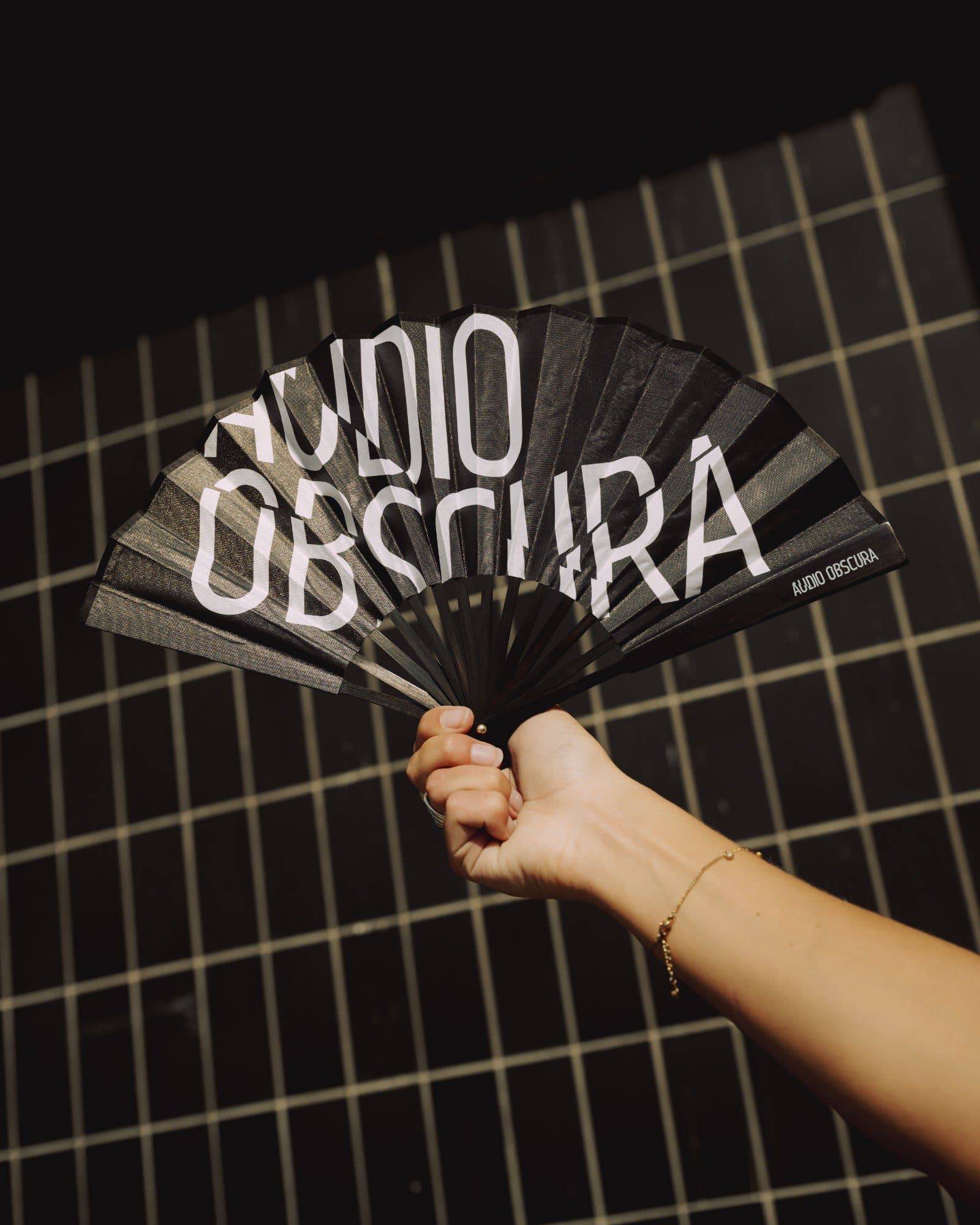 A hand holding up an open black hand fan with AUDIO OBSCURA in large white letters across the pleats and again in small letters near its outer edge, against a wall of black tiles with pale grout.