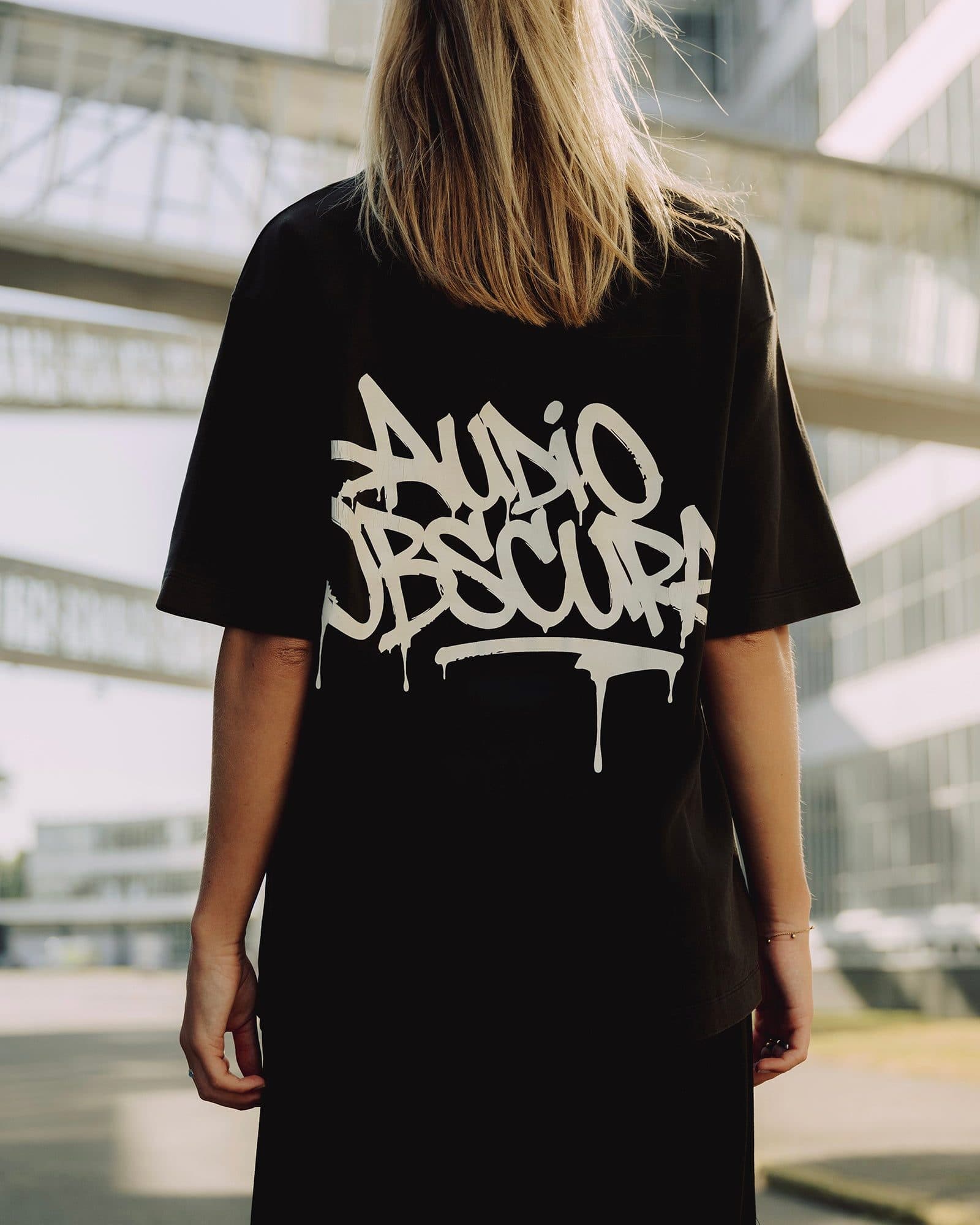 A person with long blonde hair seen from behind on a sunlit street below a glass footbridge, in a black oversized T-shirt with AUDIO OBSCURA in an off-white dripping graffiti tag across the back.