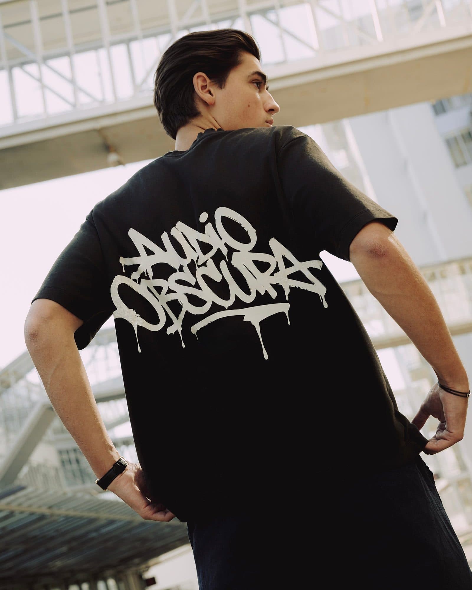 A person with dark hair seen from behind with hands on hips, looking over one shoulder below a glass footbridge, in a black T-shirt with AUDIO OBSCURA in an off-white dripping graffiti tag across the back.