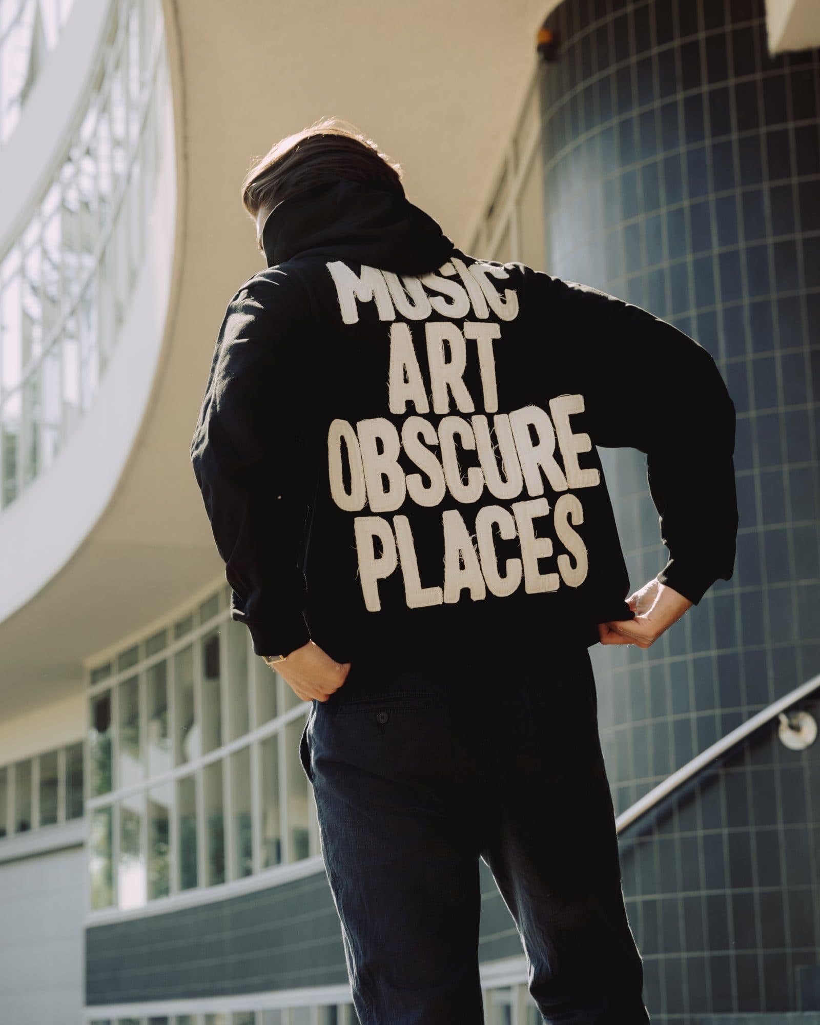 A person seen from behind with hands on hips beside a curved white building with tall windows and a tower of dark blue tiles, in a black hoodie with MUSIC ART OBSCURE PLACES in cream fabric letters with frayed edges on the back.