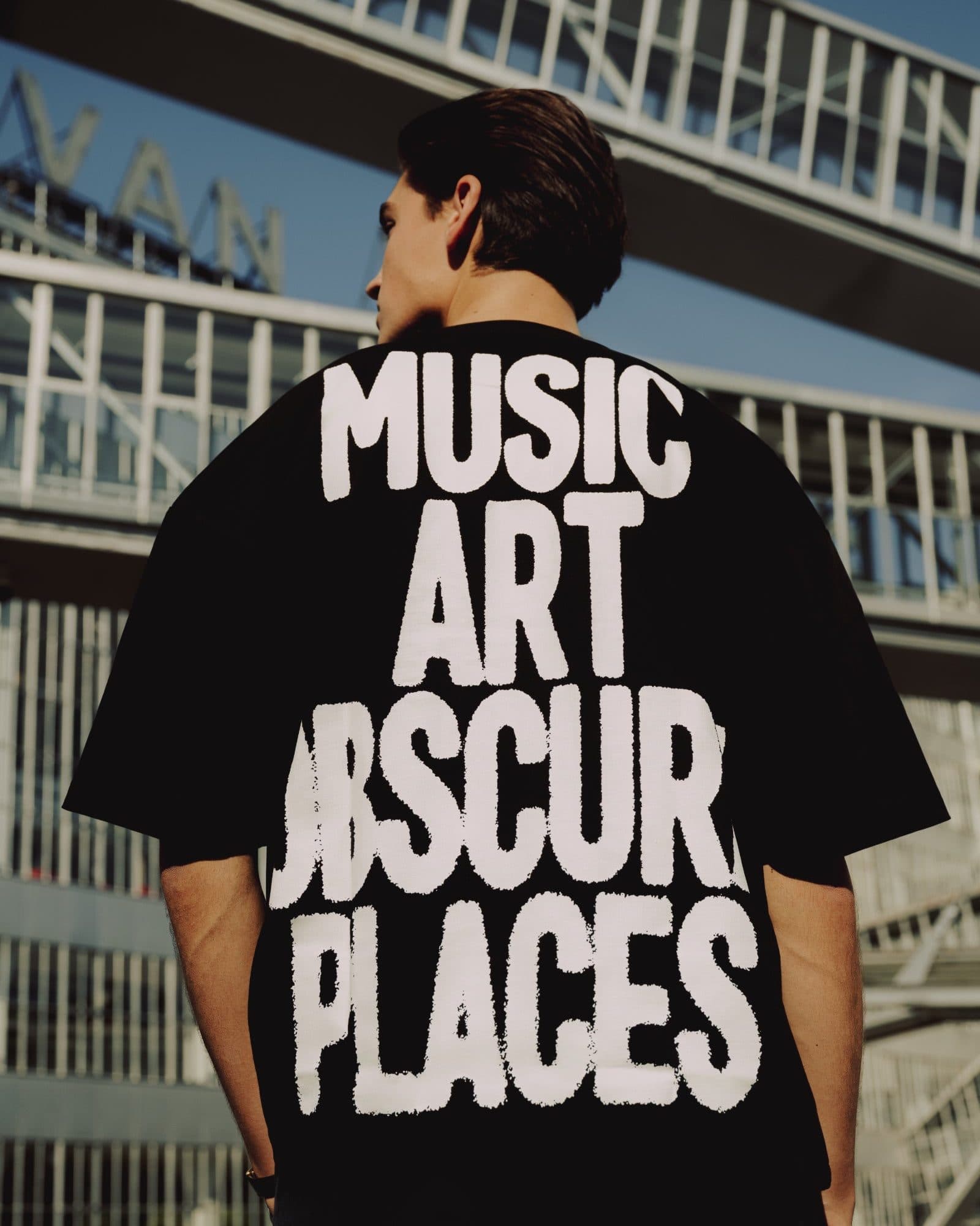 A person with dark hair seen from behind below glass footbridges against a blue sky, in a black oversized T-shirt with MUSIC ART OBSCURE PLACES in large white letters across the back, the first and last letters of OBSCURE partly hidden at the sides.