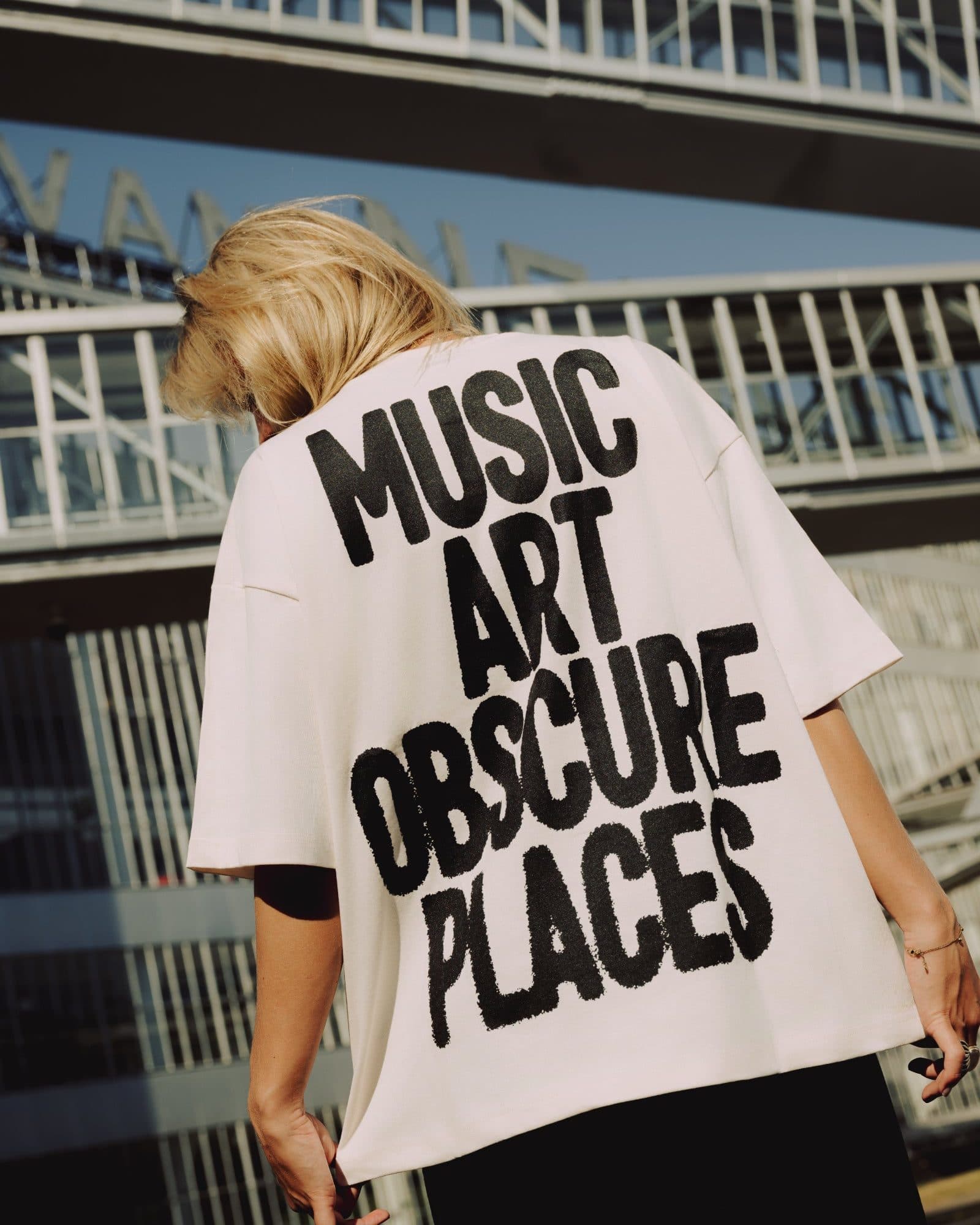 A person with blonde hair seen from behind, head bowed, below glass footbridges against a blue sky, in an off-white oversized T-shirt with MUSIC ART OBSCURE PLACES in large black letters across the back.