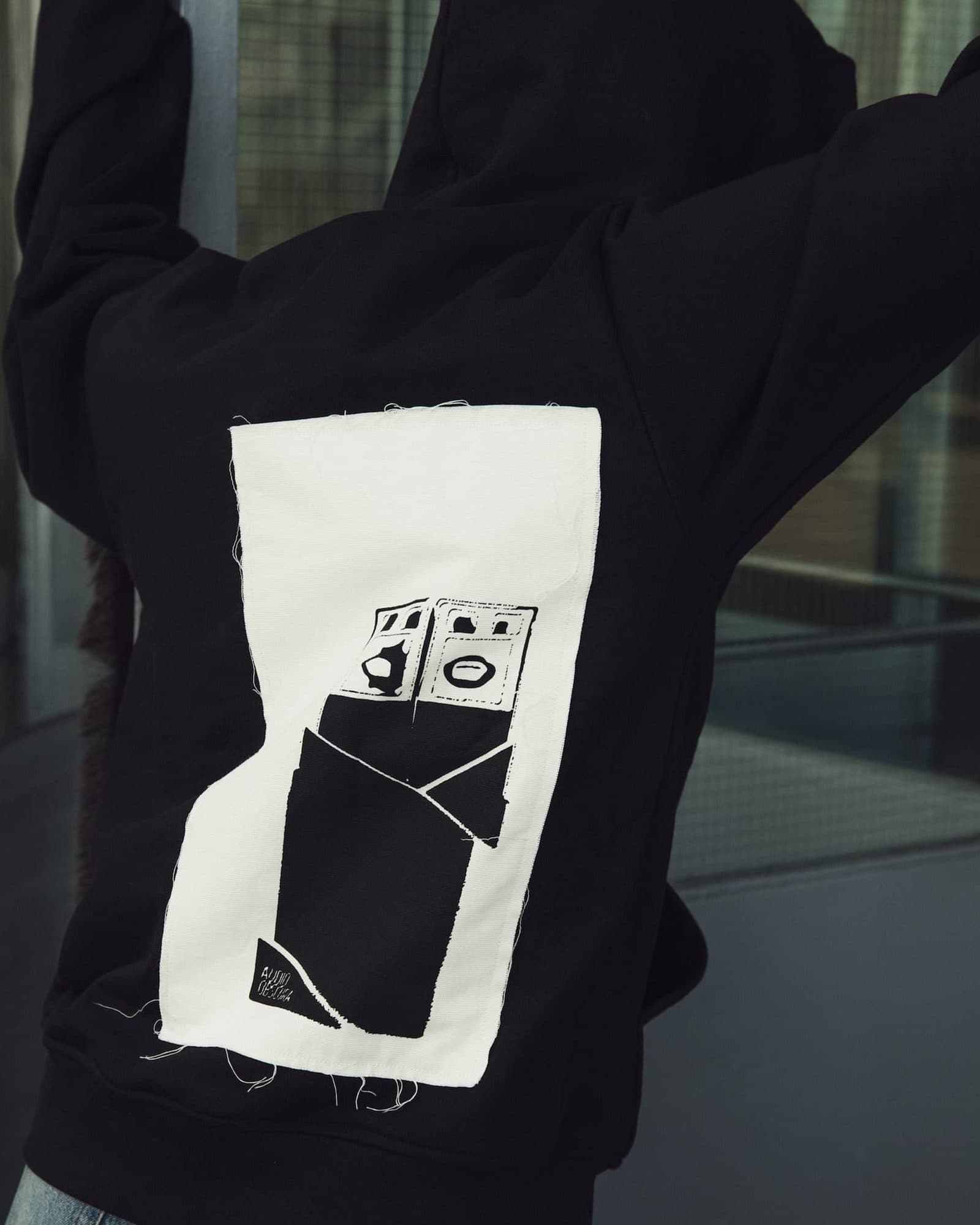 Close-up of the back of a black hoodie, worn with both arms raised in front of dark glass panes, with a cream patch with loose threads at its edges, printed in black with a drawing of a speaker stack and a small hand-lettered AUDIO OBSCURA.