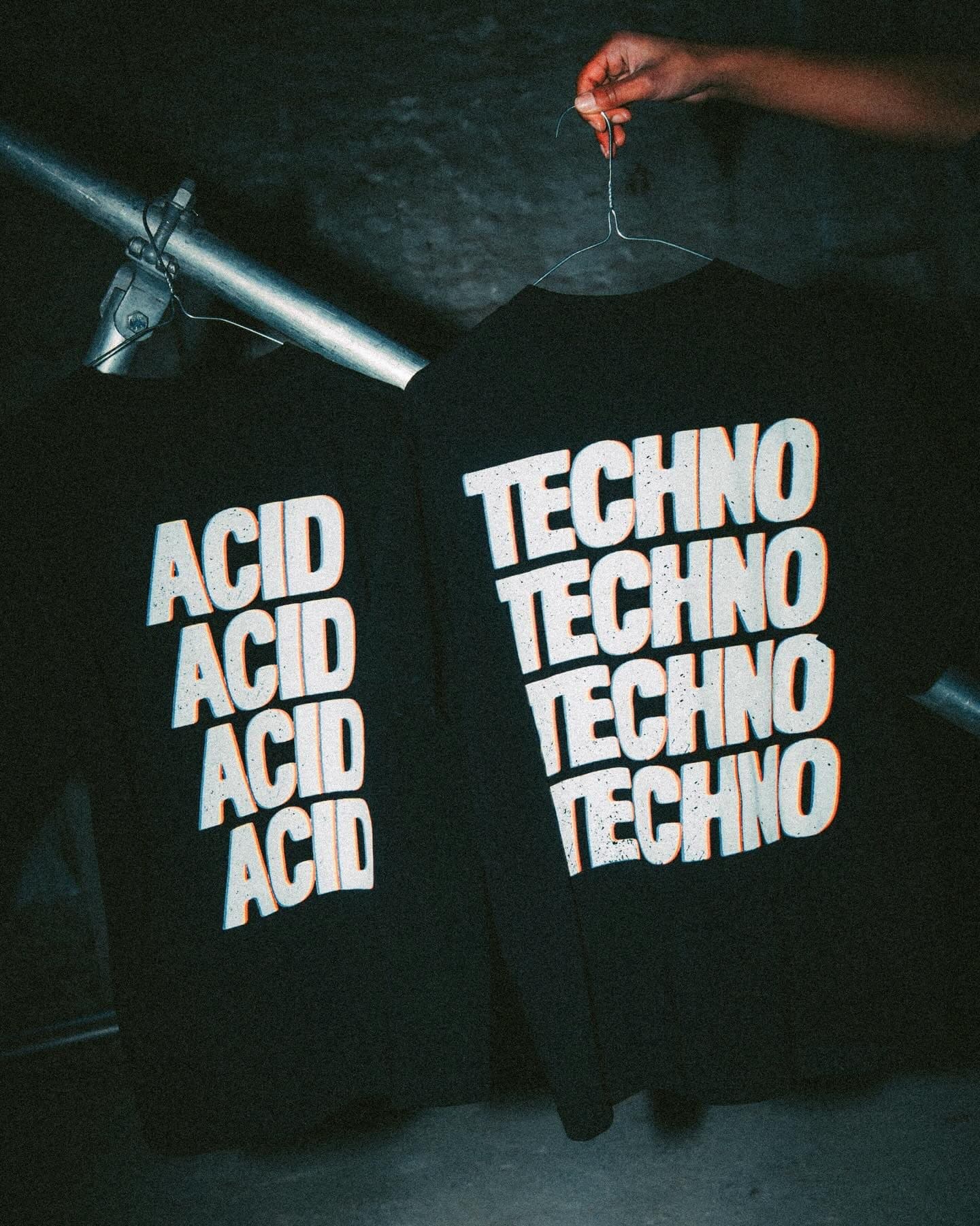 Backs of two black T-shirts on wire hangers in flash light, printed with ACID and with TECHNO four times each in large white letters edged in orange, one held up by a hand beside a metal pipe.