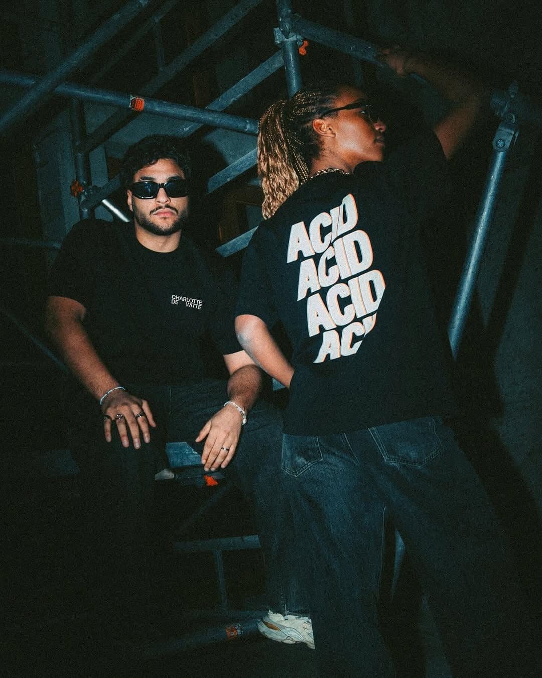 Two people among scaffolding, lit by a flash in the dark, one seated in sunglasses and a black T-shirt with CHARLOTTE DE WITTE printed small on the chest, one turned away in a black T-shirt with ACID four times in large white letters edged in orange on the back.