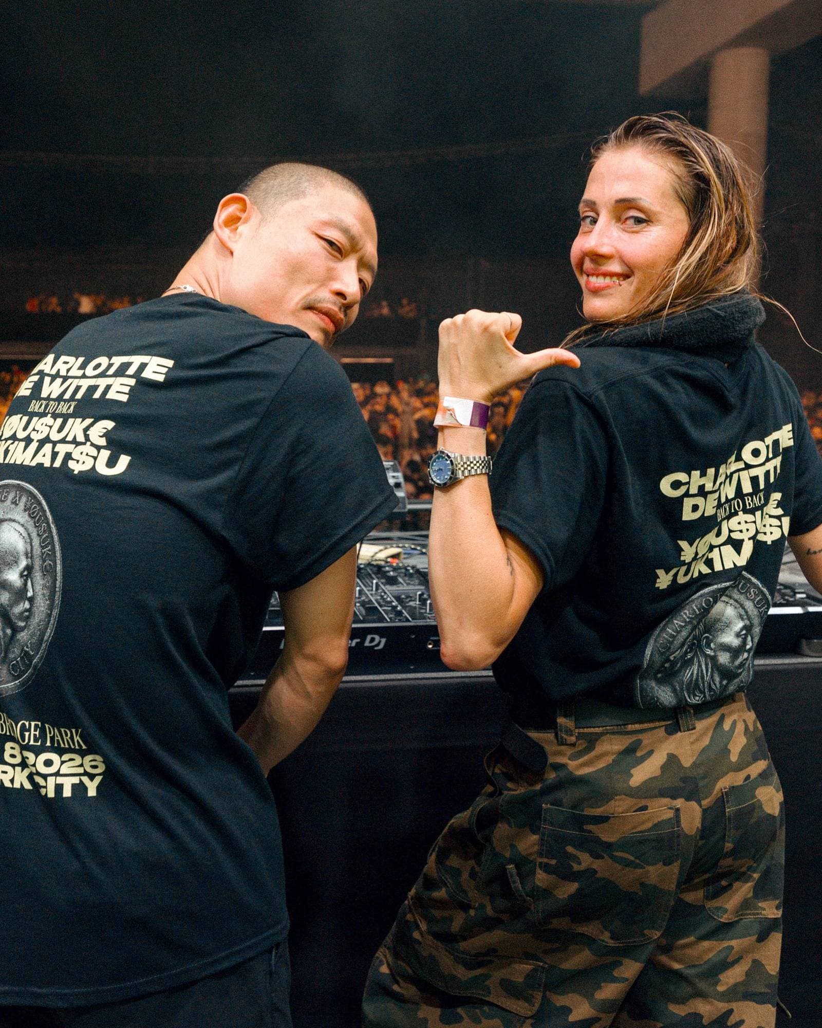 Two people at the decks in front of a packed crowd, looking back over their shoulders, one pointing a thumb, in black T-shirts with a cream back print: CHARLOTTE DE WITTE, BACK TO BACK and YOUSUKE YUKIMATSU written with yen, dollar and euro signs, a silver coin with a profile, and BRIDGE PARK and 2026 lower down.