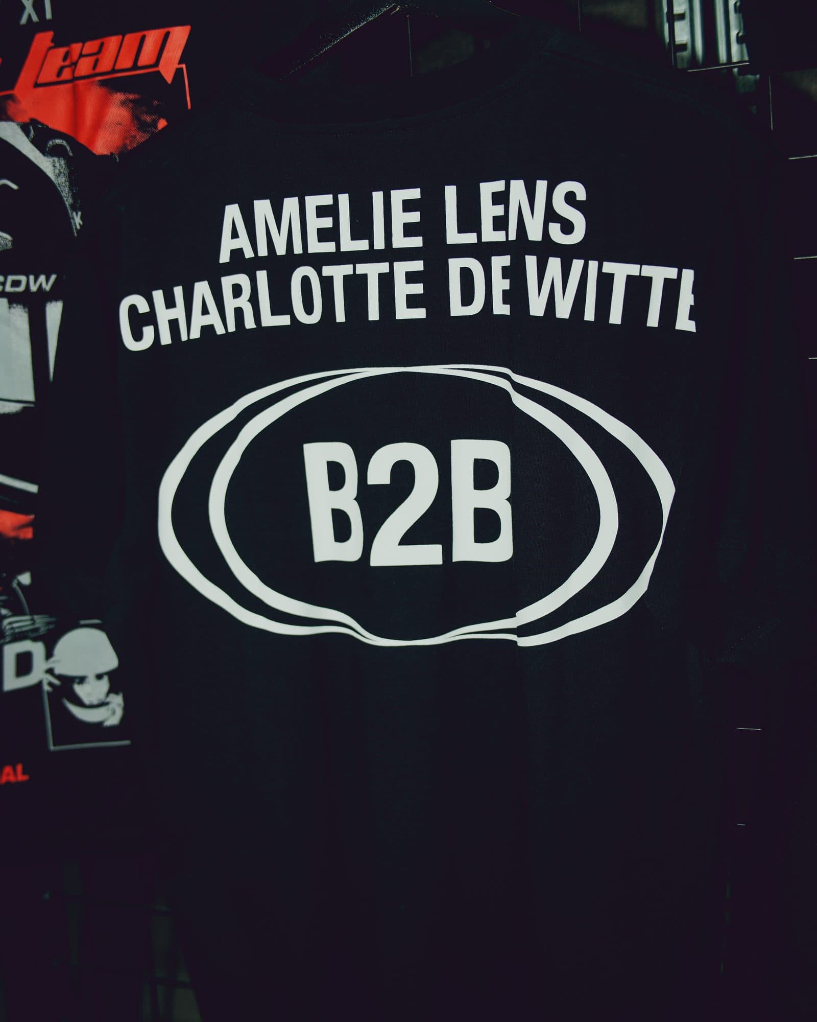 Back of a black T-shirt on a rack, printed in white with AMELIE LENS and CHARLOTTE DE WITTE above B2B inside overlapping wavy ovals, beside part of another black tee printed with a red team graphic, CDW lettering and helmet images.