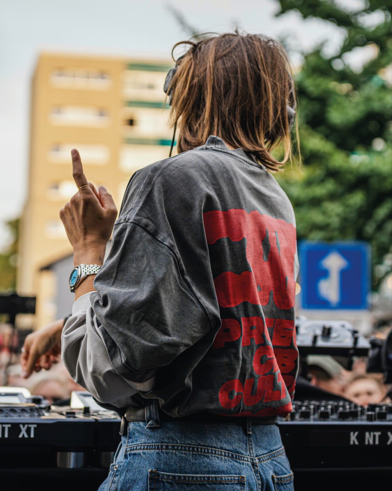 A person at the decks of a street party seen from behind, one finger raised, in a washed grey top with paler long sleeves and a large red back print in melting letters, partly hidden by an arm, with the KNTXT logo on the booth and a block of flats beyond.