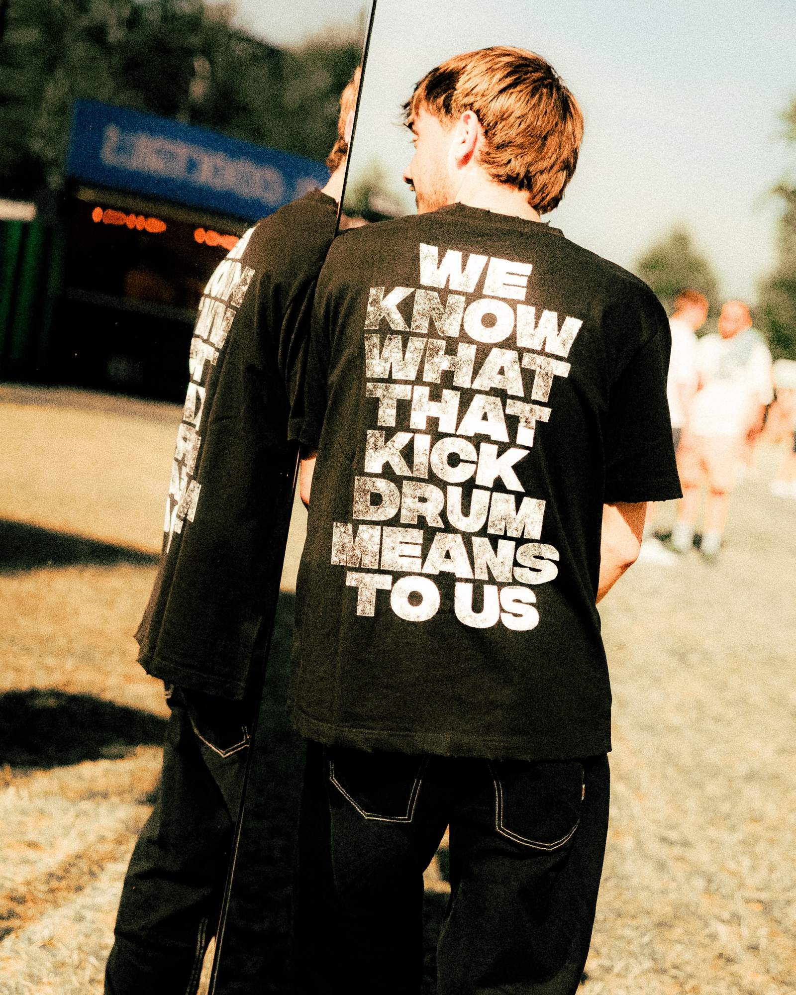 A person seen from behind beside a tall mirror on a sunny festival field, in a black T-shirt with WE KNOW WHAT THAT KICK DRUM MEANS TO US in distressed white letters across the back, the print reflected in the mirror.