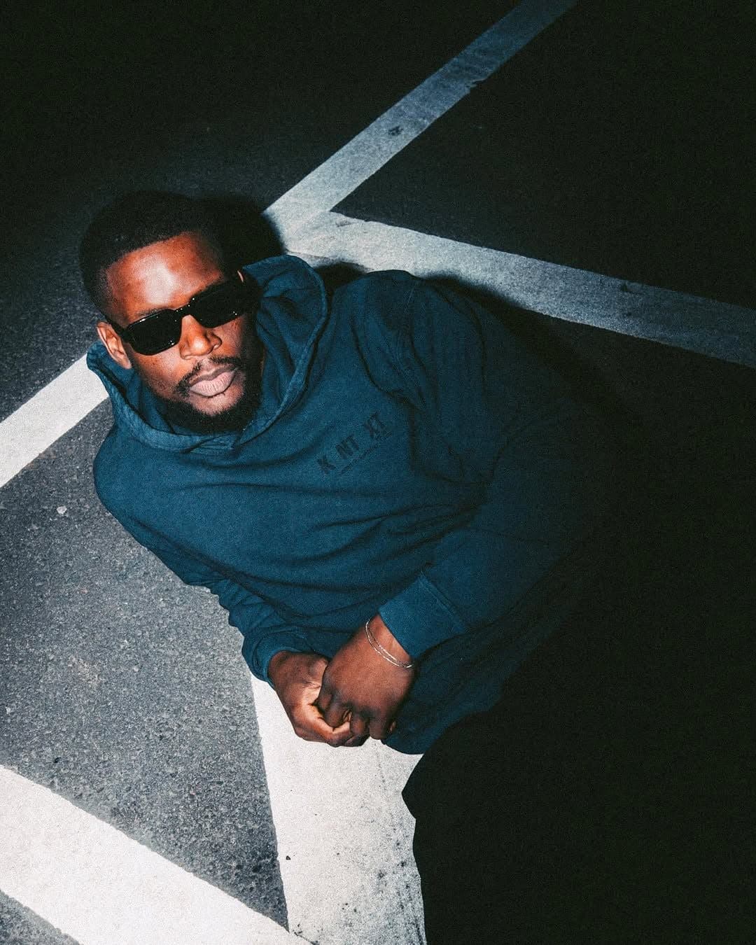 A person in sunglasses lying on asphalt with white painted lines, in a dark teal hoodie with KNTXT printed tone on tone on the chest.