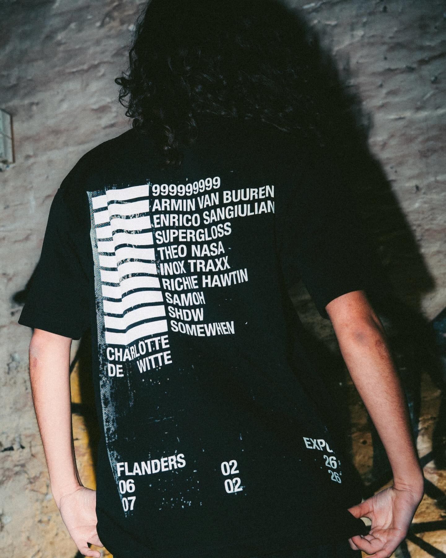 Back of a black T-shirt worn by a person with long dark curly hair against a whitewashed brick wall in flash light, printed in white with a stack of wavy bars beside a list of names, CHARLOTTE DE WITTE below them and FLANDERS, EXPO and dates near the hem.