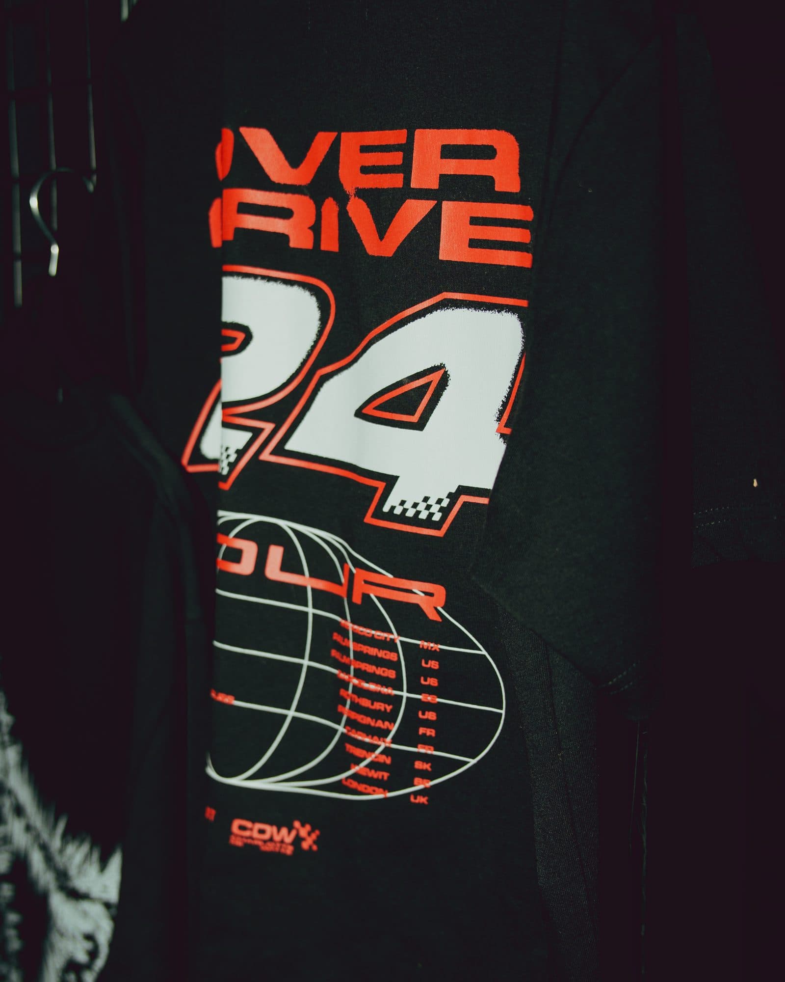 Back of a black T-shirt on a hanger, printed in red and white with OVERDRIVE, its first letters hidden by a garment in front, a large 24, TOUR, a wireframe globe with a list of tour cities and a CDW logo.