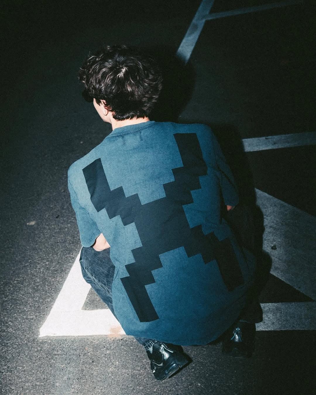 A person crouching on asphalt with white painted lines, lit by a flash in the dark, seen from behind in a washed blue T-shirt with a large black pixelated X across the back.