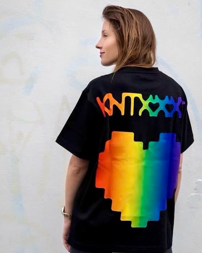A person in profile against a white wall, seen from behind in a black T-shirt with a rainbow back print: KNTXXX in dripping letters, with heart-shaped gaps between the X's, above a large pixelated heart.