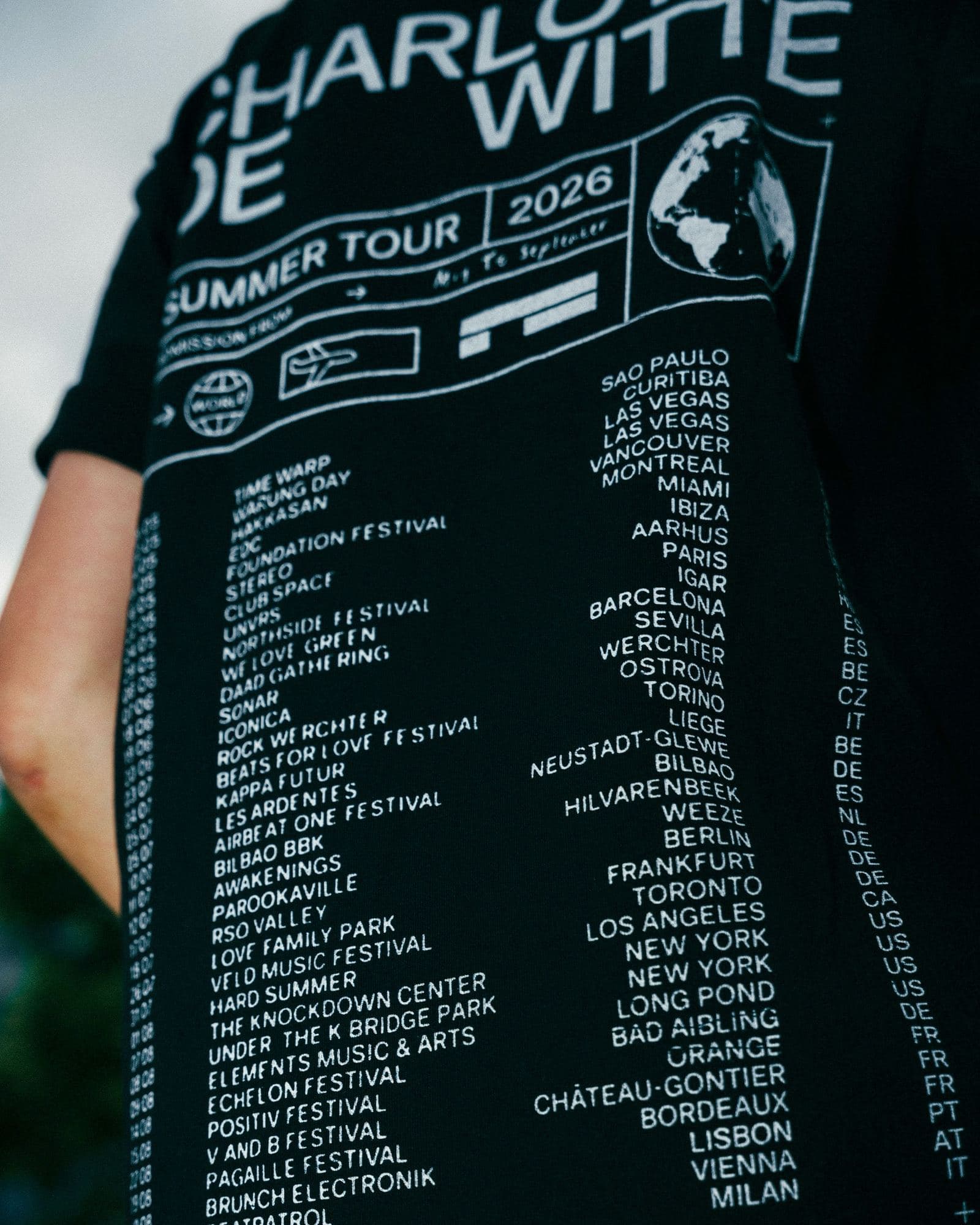 Close-up of the back of a black T-shirt printed in white with CHARLOTTE DE WITTE, SUMMER TOUR 2026, a globe, small travel icons and a long list of dates, festivals, cities and country codes.