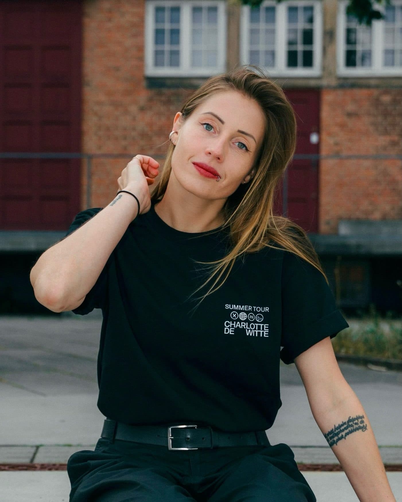 A person sitting on concrete steps in front of a brick building, one hand in the hair, in a black T-shirt with SUMMER TOUR, four small round icons and CHARLOTTE DE WITTE printed in white on the chest.