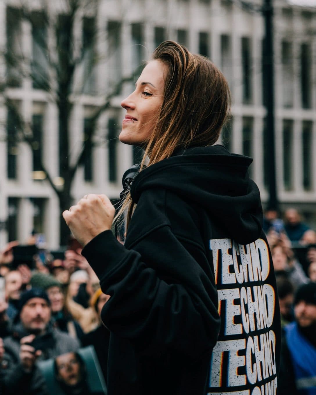 A person in profile raising a fist above a crowd in a city square, in a black hoodie with TECHNO repeated in large white letters down the back, the print running off the edge.