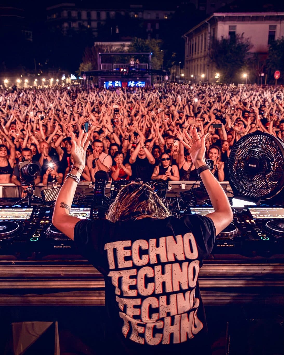A person at the decks seen from behind with both arms raised to a packed open-air crowd at night, in a black T-shirt with TECHNO four times in large pale letters edged in orange on the back.
