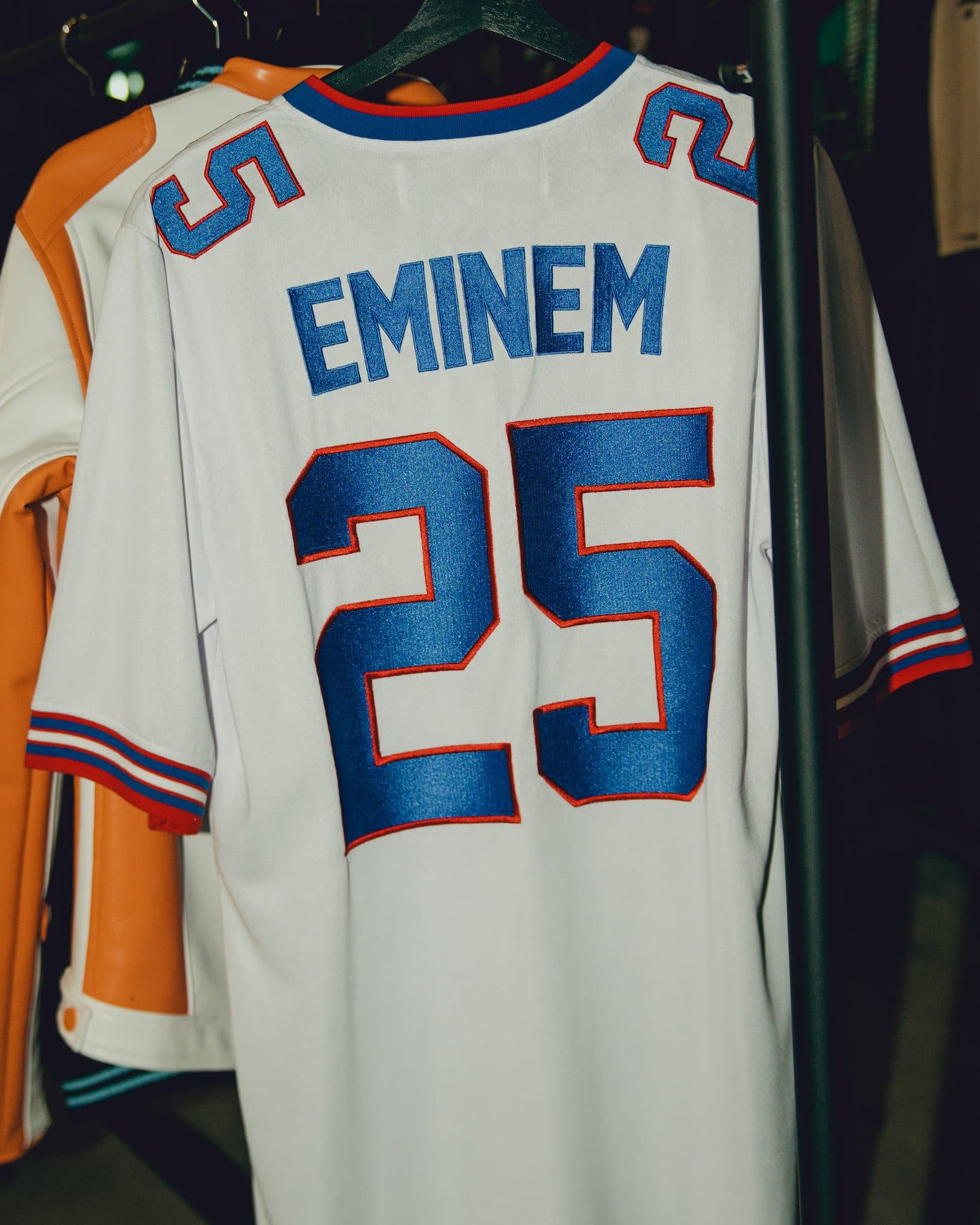 Back of a white football jersey with EMINEM and a large 25 in blue outlined in red, numbers on the shoulders and striped cuffs, on a hanger on a clothes rail beside an orange and white jacket.
