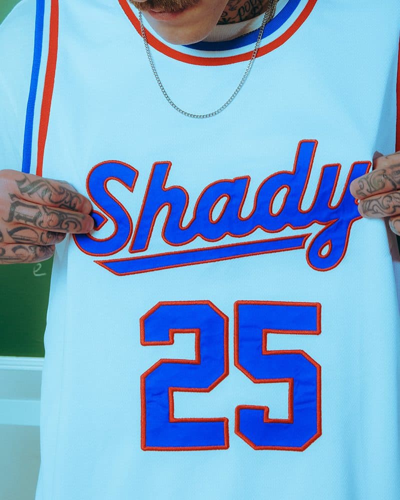 A person with tattooed hands pulling forward a white sleeveless mesh jersey worn over a white T-shirt, with Shady in blue script outlined in red above a large blue 25, blue and red striped trim at the collar and armholes, a silver chain above it.