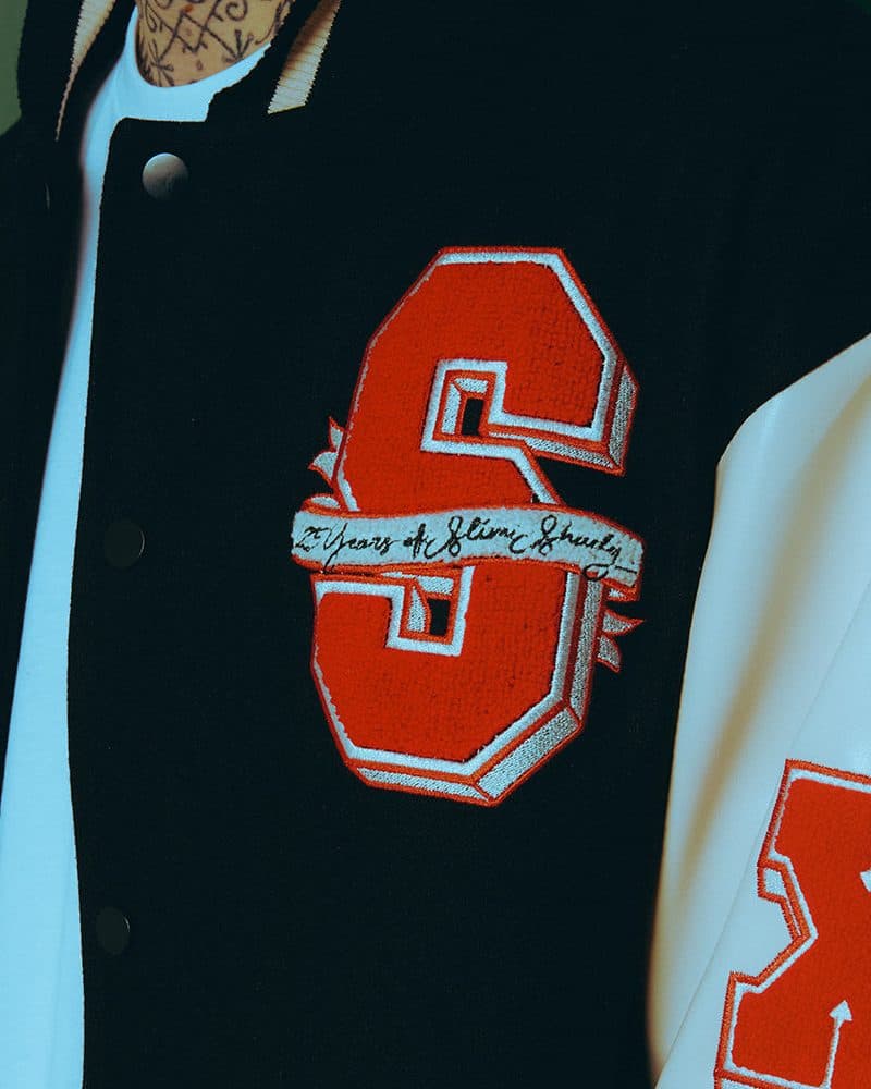 Close-up of the red chenille S patch edged in white on the black chest of the Slim Shady 25 varsity jacket, with a cream banner reading 25 Years of Slim Shady in black script across it, snap buttons, a white T-shirt underneath and part of a red X on the cream sleeve.