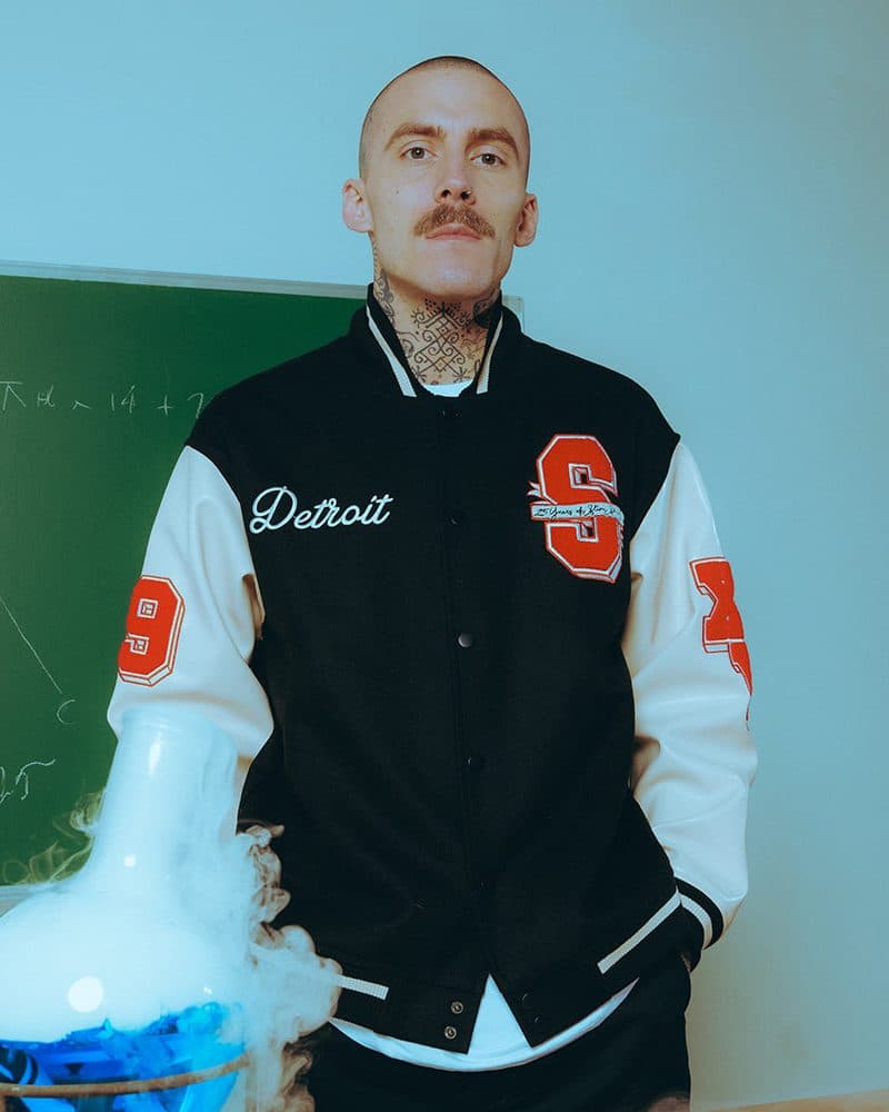 The Slim Shady 25 varsity jacket worn by a person with neck tattoos: black with cream sleeves, Detroit in white script on the chest, a red S patch with a banner on the other side and a red 9 and X on the sleeves, beside a smoking glass flask in front of a blackboard.