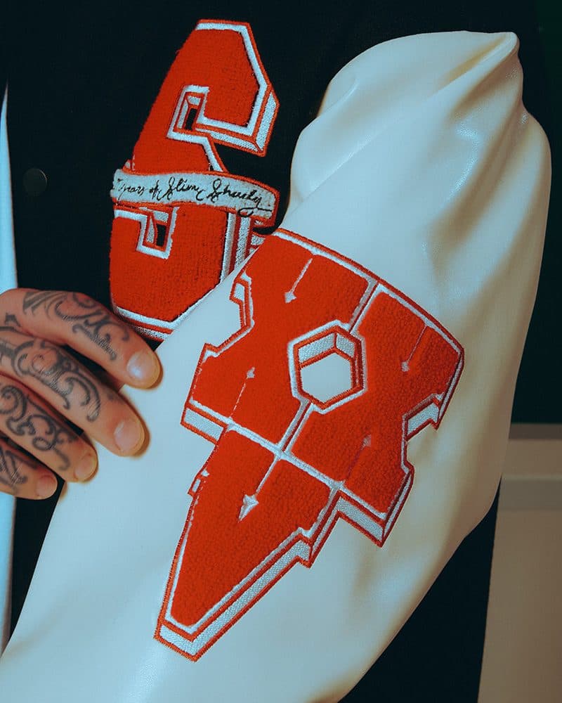 A tattooed hand beside the cream sleeve of the Slim Shady 25 varsity jacket with its red chenille X patch edged in white, a hexagon at the centre and a long point below, and behind it, on the black chest, part of the red S patch with its script banner.