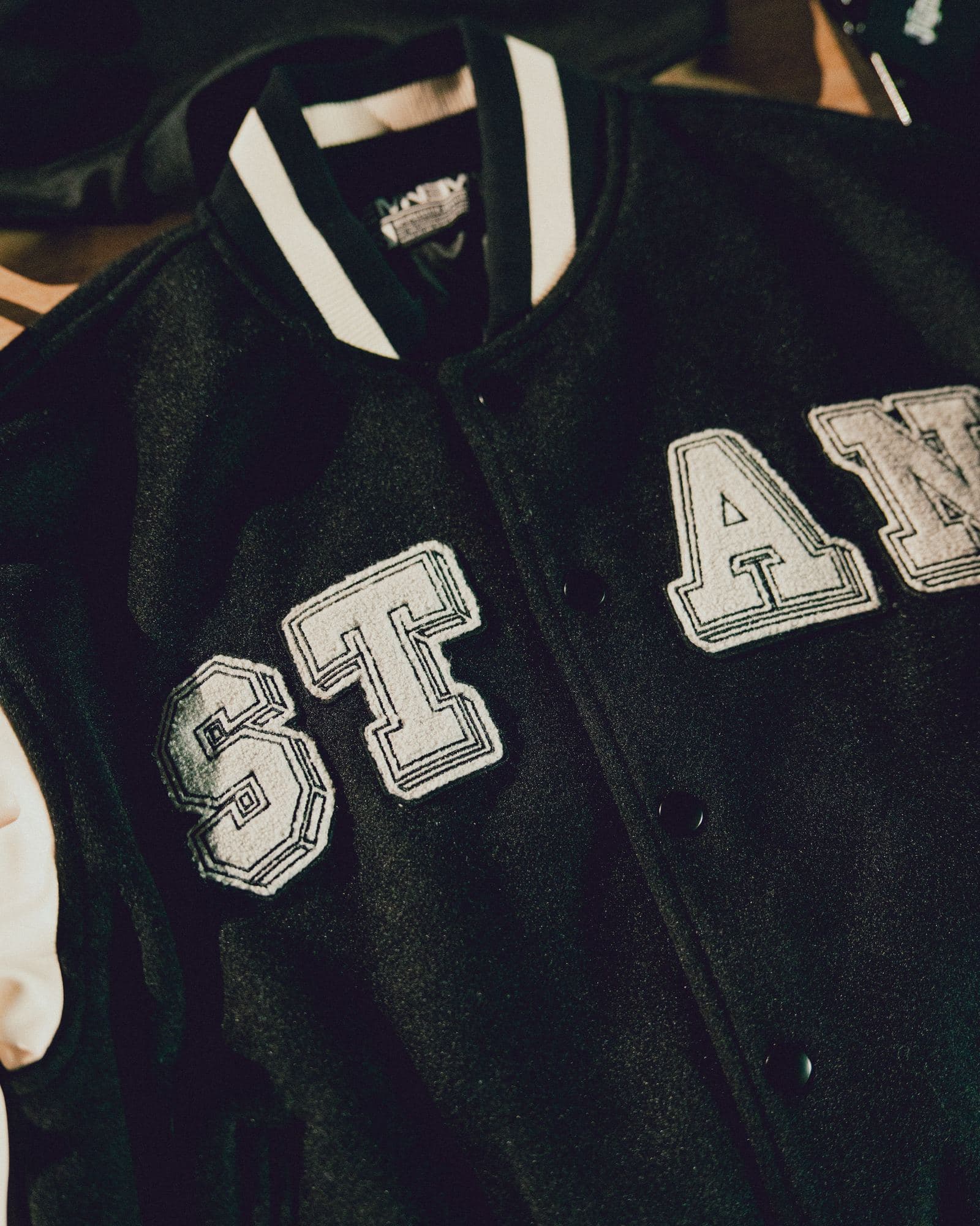Front of a black varsity jacket with a cream sleeve, cream chenille letters spelling STAN across the chest, a black and white striped rib collar, black snap buttons and a label inside the collar.
