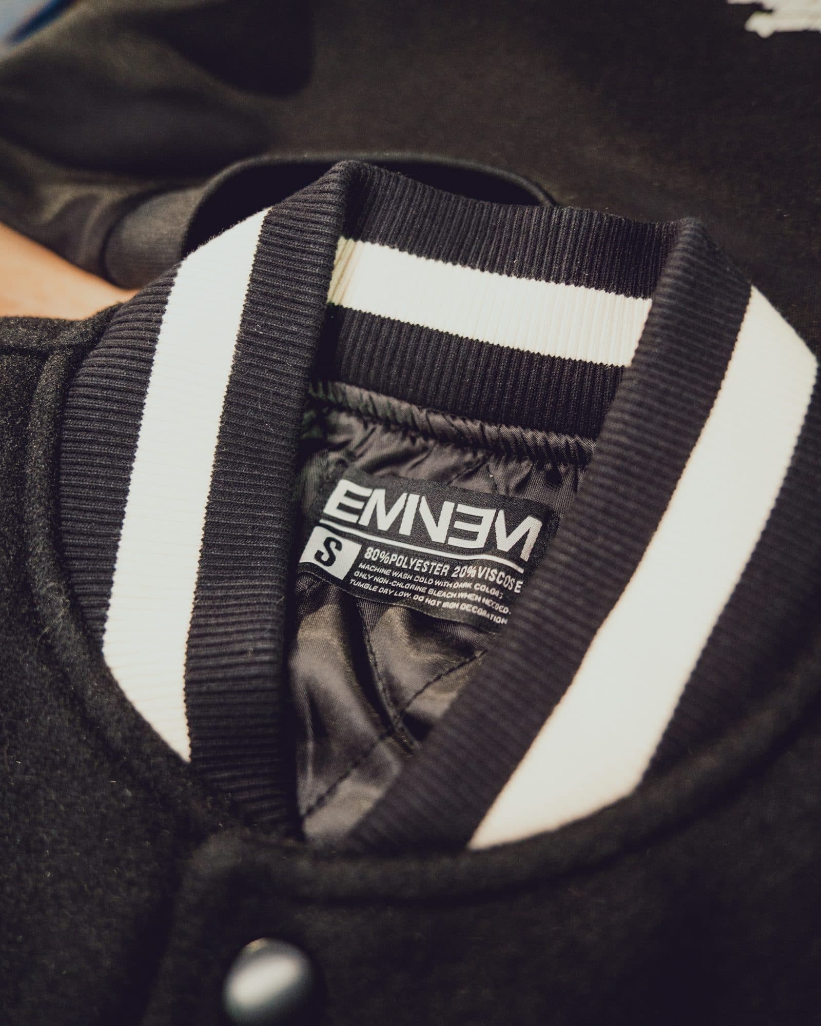 Collar of a black varsity jacket with black and white striped ribbing, a shiny black lining and a black Eminem label with size S and care text.