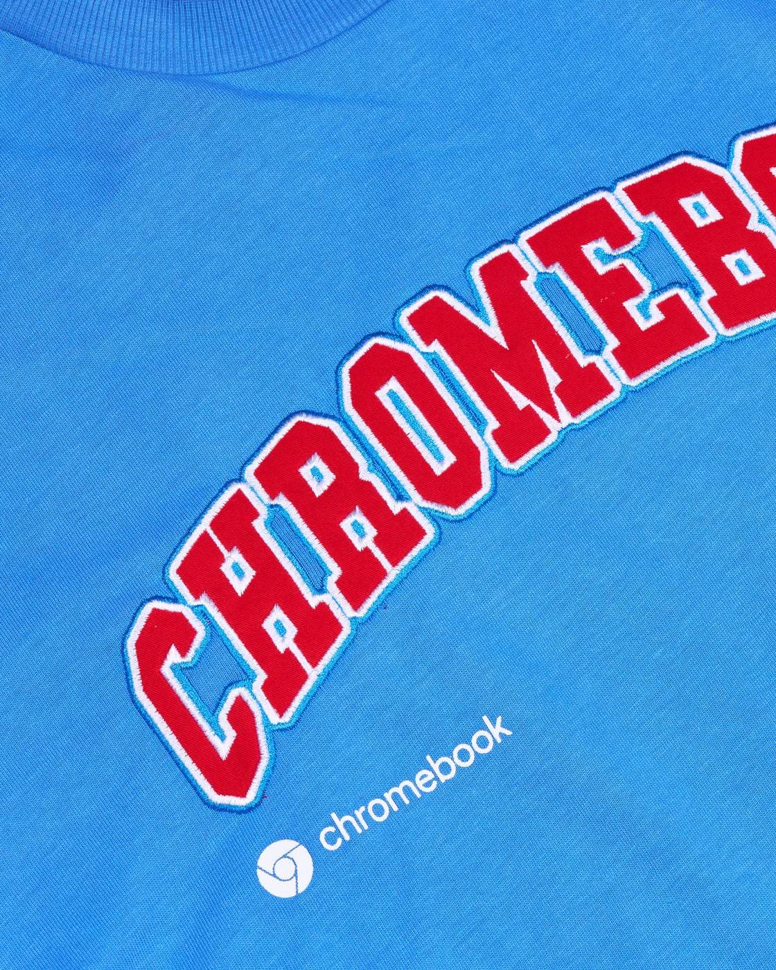 Blue crewneck with an arch of red appliqué letters, edged in white and light blue stitching, that runs off the right edge after CHROMEB, above a small white printed chromebook logo.